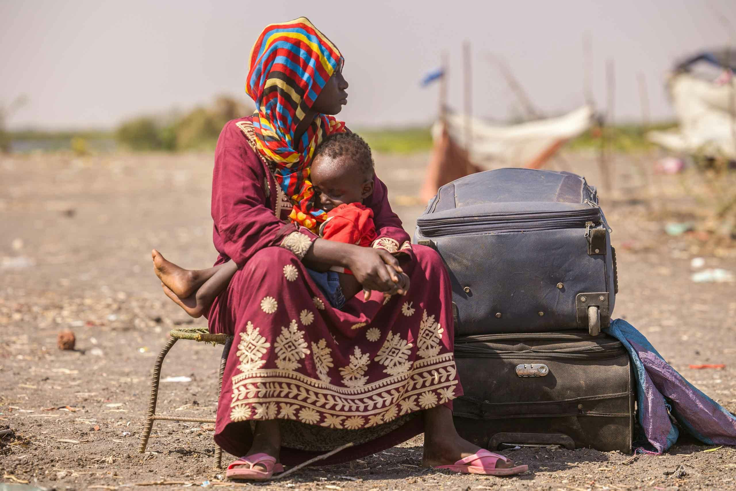sudanesische Frau mit einem Kleinkind sitzt inmitten einer kargen Landschaft neben 2 Koffern