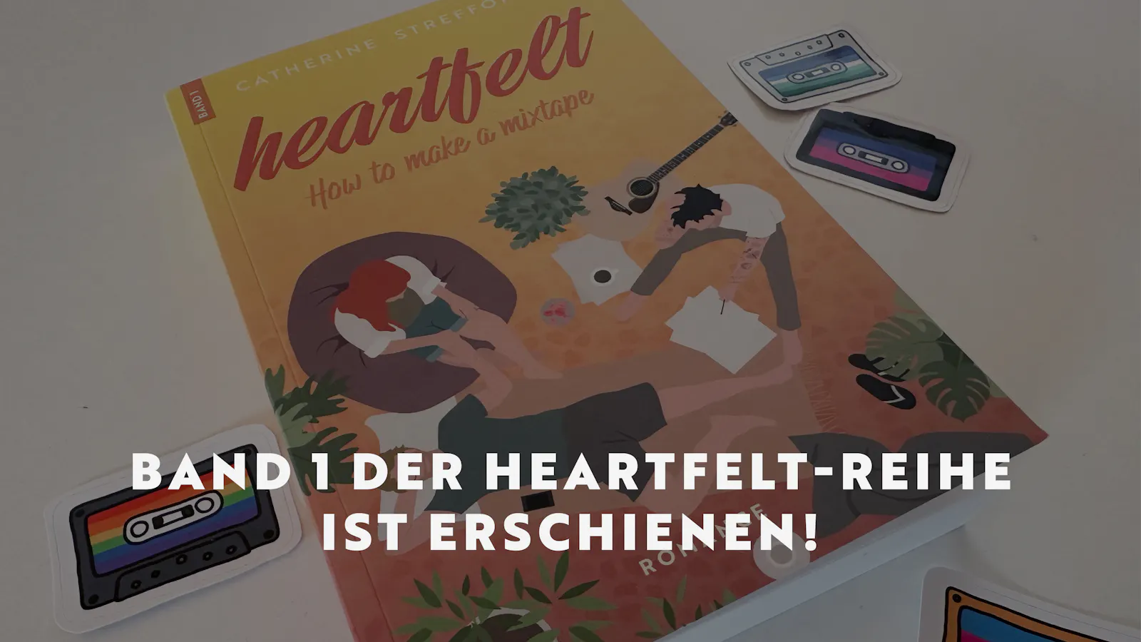 Das Taschenbuch von Heartfelt Band 1 liegt auf einem weißen Tisch, darum verteilt liegen Aufkleber von Musikkassetten in den Farben verschiedener Pride-Flaggen.