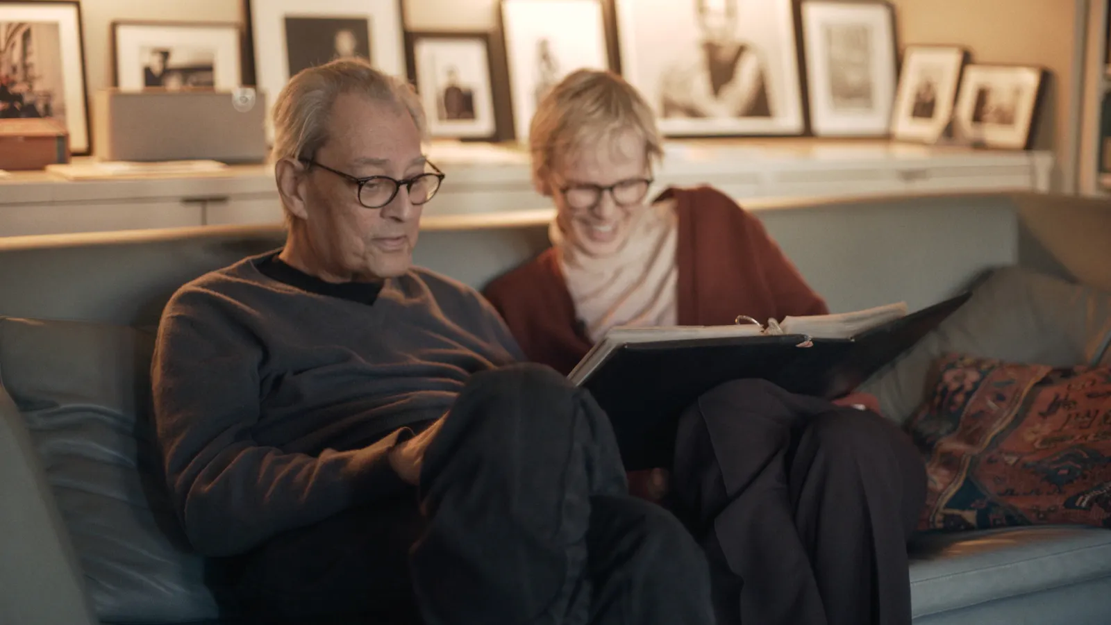 Filmstill aus SIRI HUSTVEDT – DANCE AROUND THE SELF: Paul Auster und Siri Hustvedt sitzen gemeinsam auf einer Couch, beide mit Brille. Auster hält ein Buch. Im Hintergrund gerahmte Fotografien an der Wand. Warmes, häusliches Licht. | Foto: IFB/Medea Film Factory