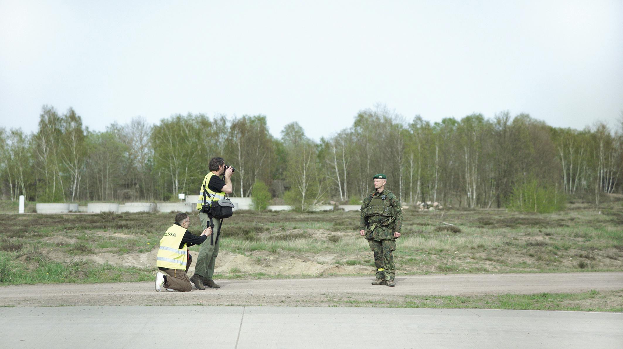 Zwei Kameraleute in gelben Sicherheitswesten mit "MEDIA"-Aufschrift filmen einen einzelnen Bundeswehrsoldaten in Uniform, der vor ihnen auf asphaltiertem Gelände steht. Im Hintergrund erstreckt sich kahles Übungsgelände mit Birkenwald. Filmstill aus SZENARIO von Marie Wilke.
