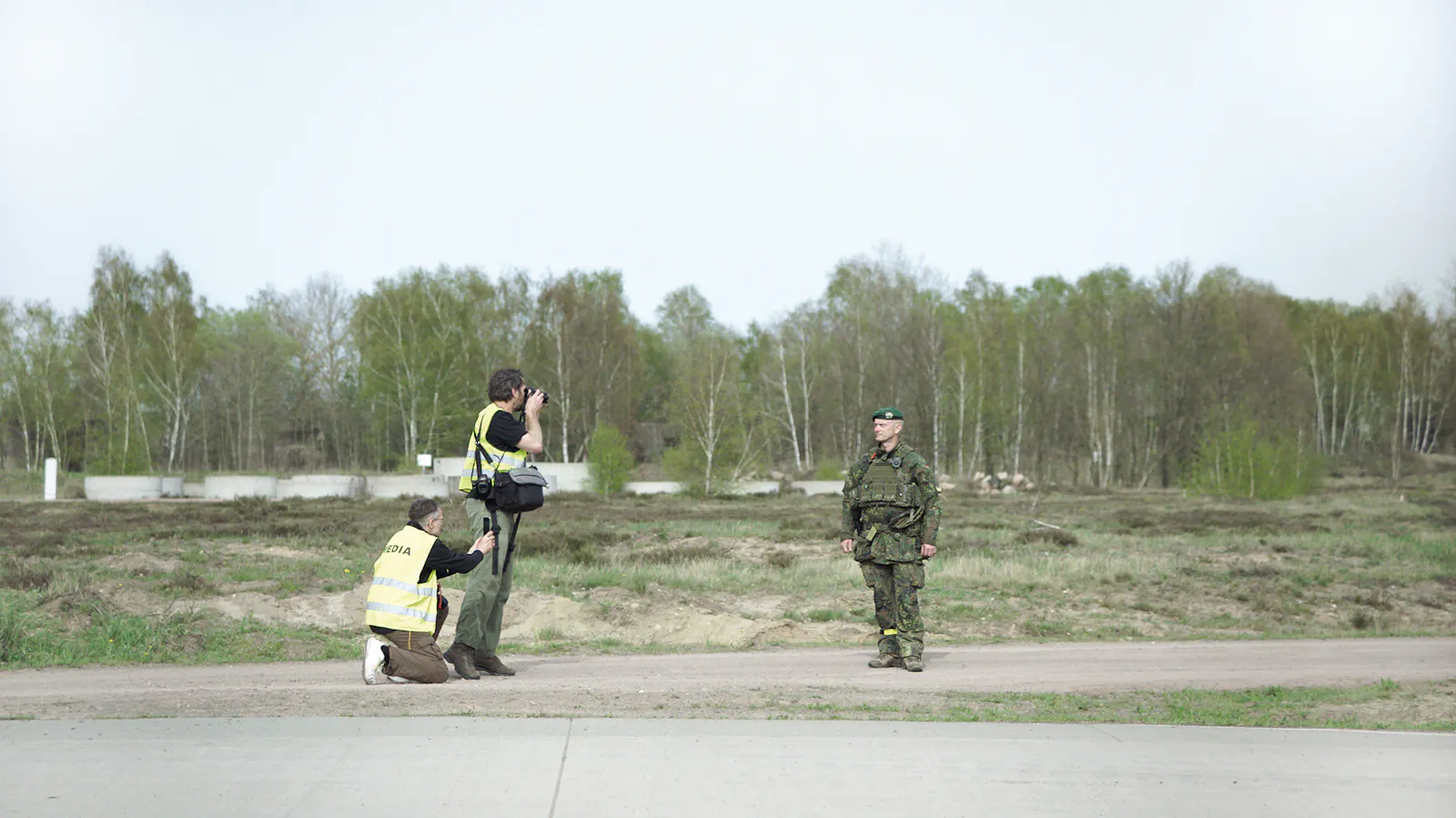 Zwei Kameraleute in gelben Sicherheitswesten mit "MEDIA"-Aufschrift filmen einen einzelnen Bundeswehrsoldaten in Uniform, der vor ihnen auf asphaltiertem Gelände steht. Im Hintergrund erstreckt sich kahles Übungsgelände mit Birkenwald. Filmstill aus SZENARIO von Marie Wilke.