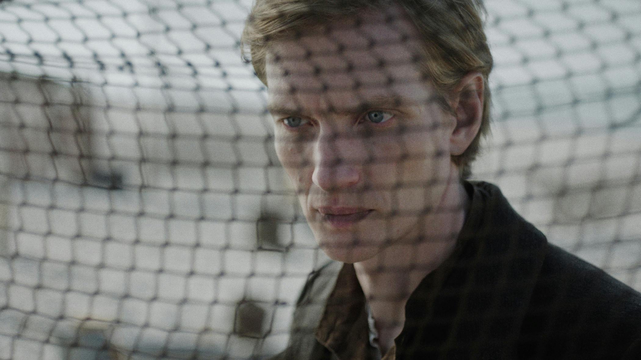 Filmstill aus DER HEIMATLOSE: Nahaufnahme eines jungen Mannes, Hein, mit mittellangen blonden Haaren und hellen Augen. Im Bildvordergrund unscharf ein Fischernetz. Er trägt dunkle Kleidung. Foto: IFB/Florian Mag.