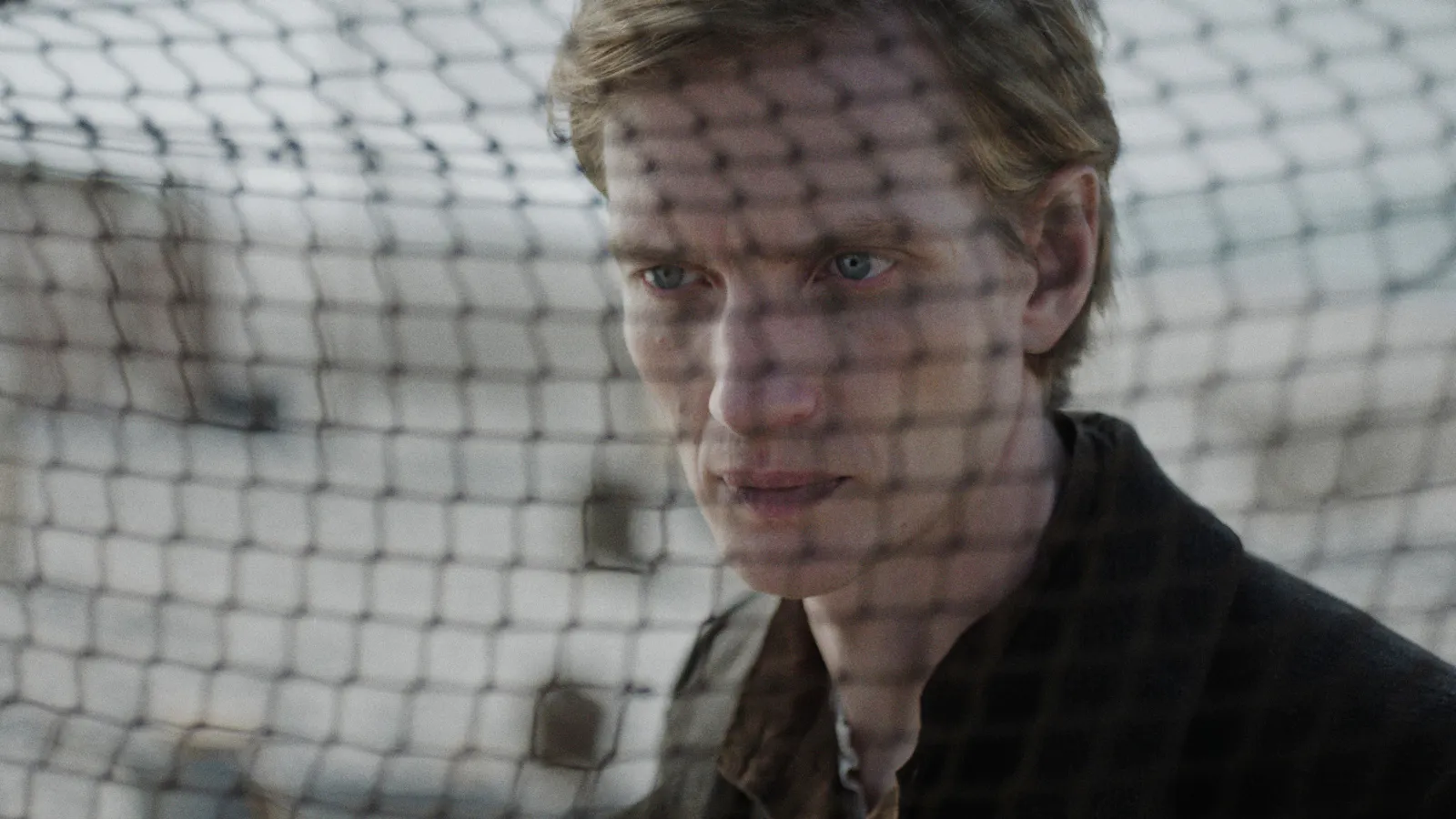 Filmstill aus DER HEIMATLOSE: Nahaufnahme eines jungen Mannes, Hein, mit mittellangen blonden Haaren und hellen Augen. Im Bildvordergrund unscharf ein Fischernetz. Er trägt dunkle Kleidung. Foto: IFB/Florian Mag.