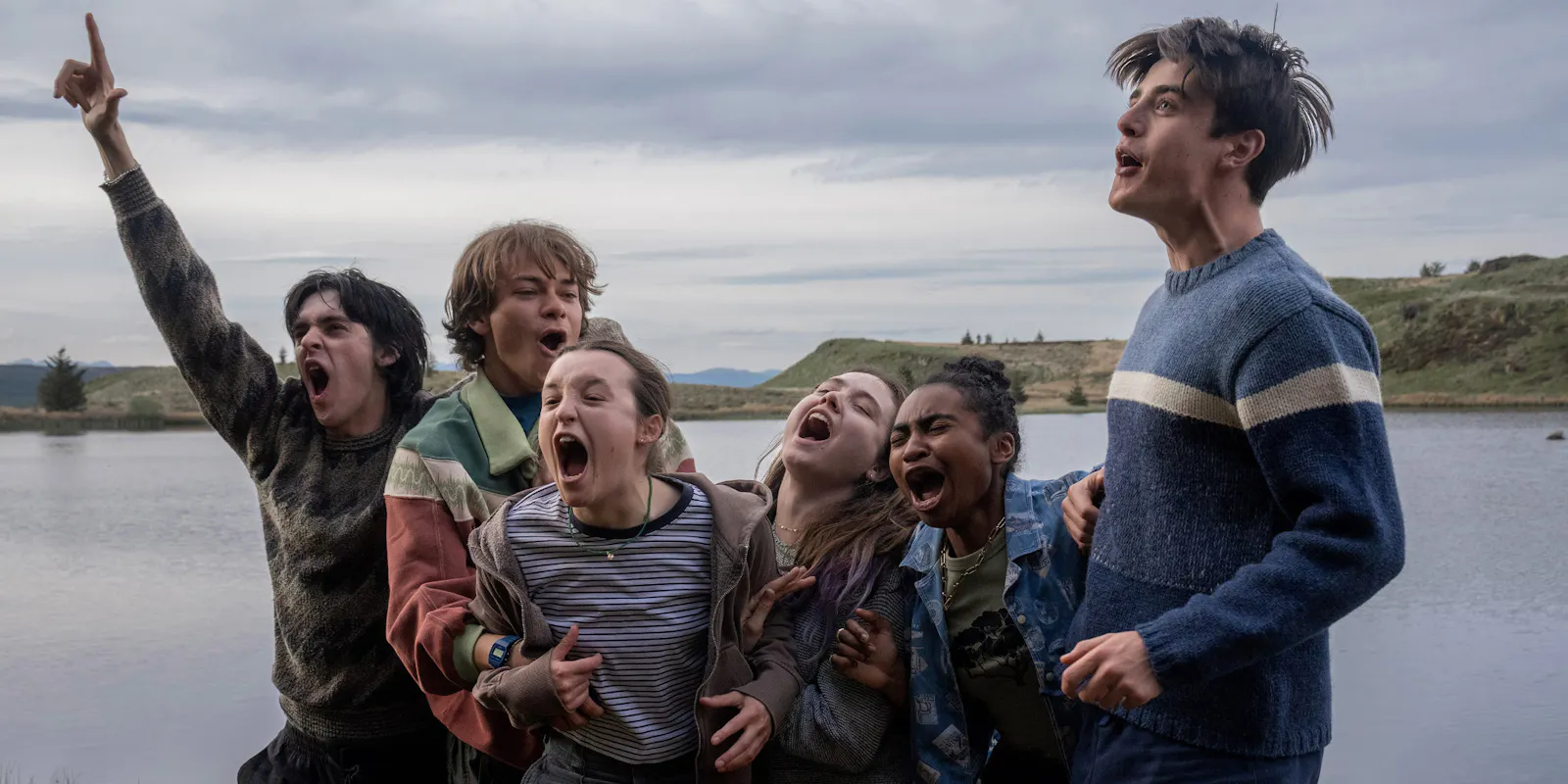 Filmstill aus SUNNY DANCER: Sechs Jugendliche stehen eng umschlungen am Ufer eines schottischen Sees, alle schreien gemeinsam in den Himmel, eine Person reckt die Faust nach oben. Hügelige Landschaft im Hintergrund unter bewölktem Himmel. Pure jugendliche Energie und Zusammengehörigkeit. Foto: IFB/Colin J Smith/Sunny Dancer Dist. Ltd.