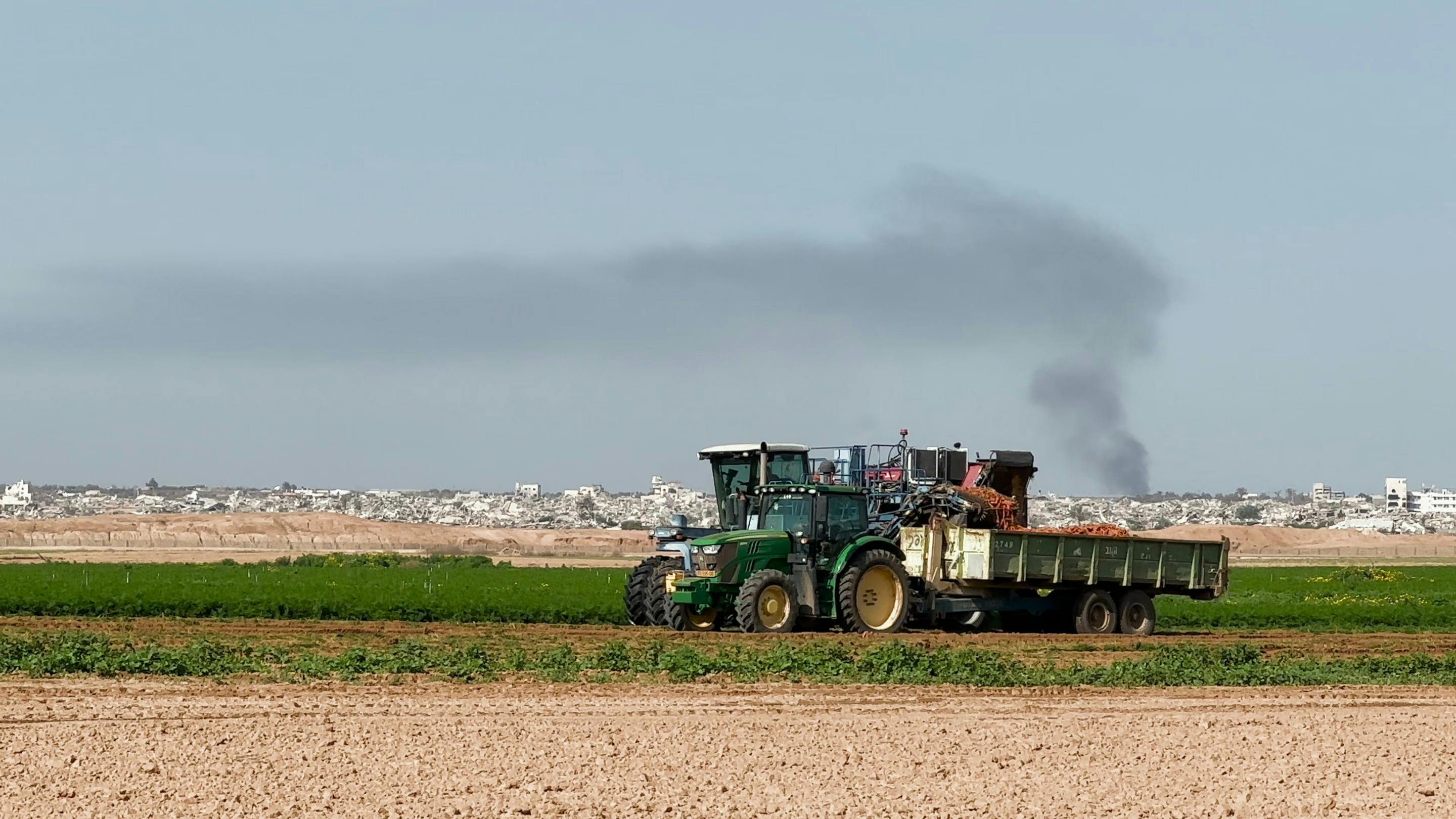 Filmstill aus EFFONDREMENT: Ein grüner Traktor mit Anhänger auf einem Feld im Vordergrund. Dahinter erstrecken sich die Trümmer von Gaza, eine dunkle Rauchsäule steigt in den Himmel. Kontrast zwischen landwirtschaftlicher Normalität und Zerstörung.