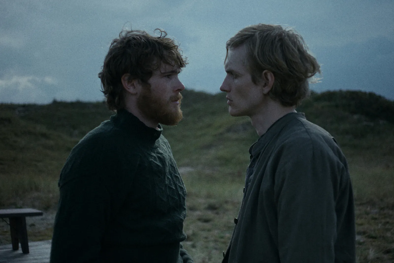 Filmstill aus DER HEIMATLOSE: Zwei Männer stehen sich in der Dämmerung direkt gegenüber und blicken sich in die Augen. Links ein Mann mit dunklen lockigen roten Haaren und Bart (Friedemann), rechts ein blonder Mann (Hein). Beide in dunkler, einfacher Kleidung. Dünenlandschaft im Hintergrund. Foto: IFB/Florian Mag.