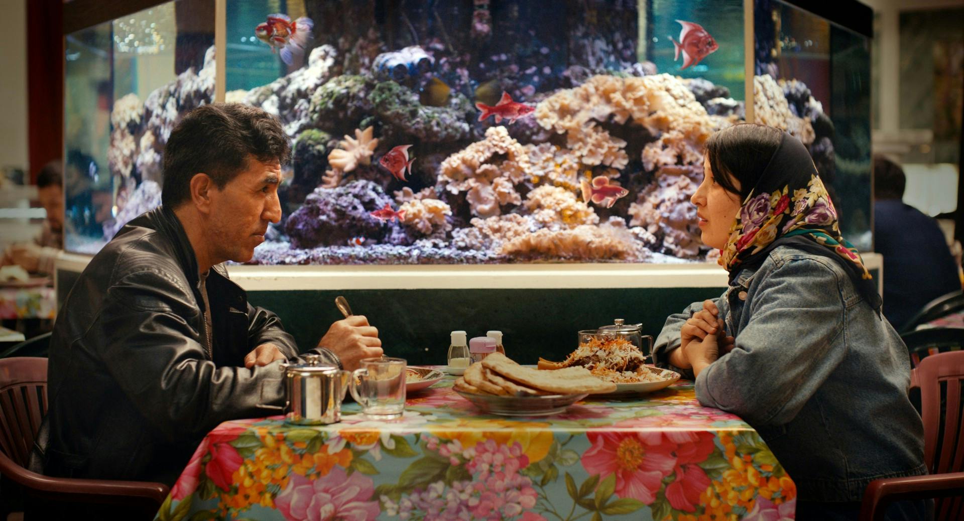 Filmstill aus NO GOOD MEN: Naru (rechts) und ein männlicher Kollege sitzen sich an einem Restaurant-Tisch gegenüber, vor ihnen Teller mit Essen auf einer bunt geblümten Plastik-ischdecke. Im Hintergrund ein großes Salzwasseraquarium mit Korallen und tropischen Fischen.