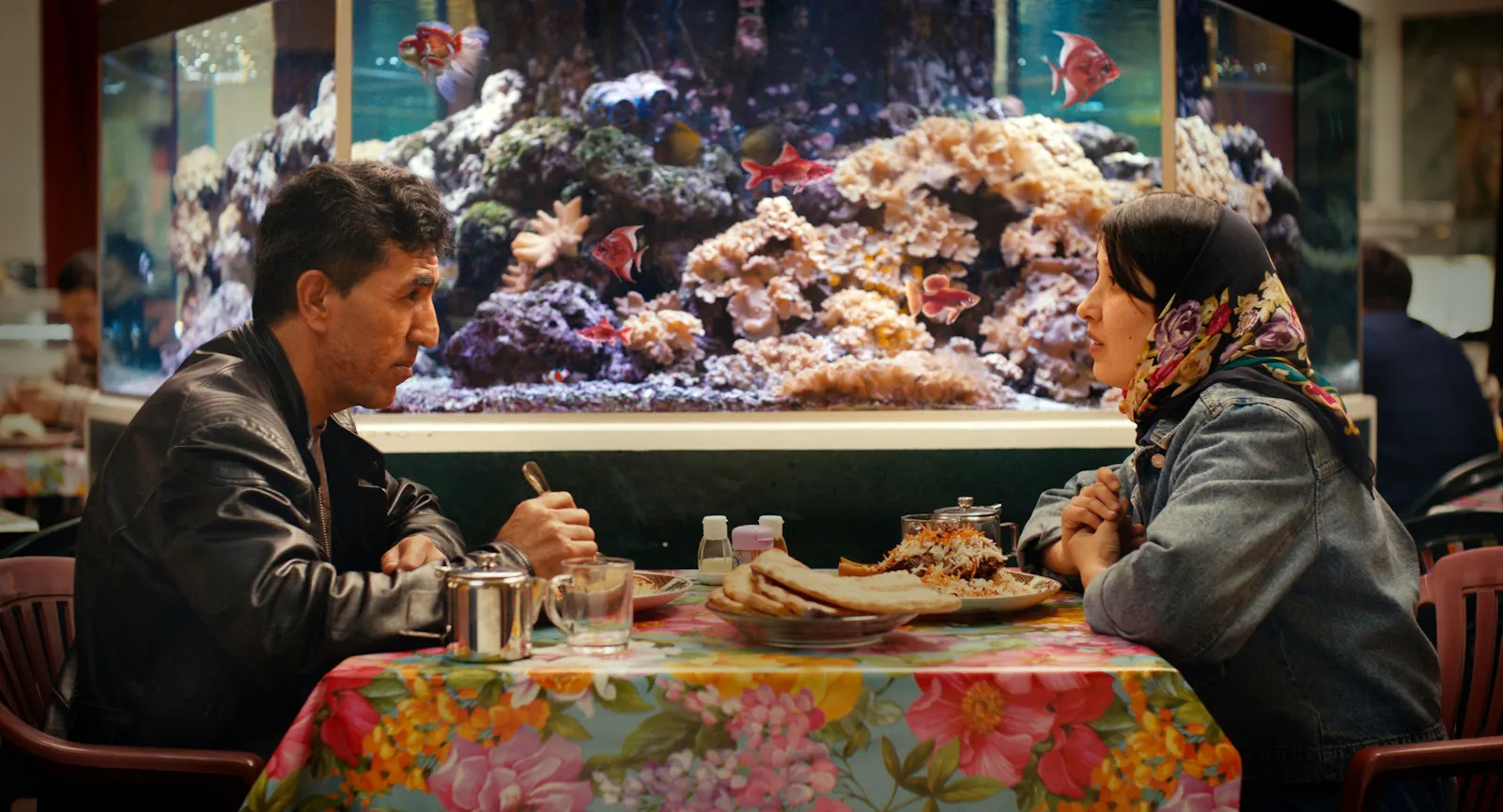 Filmstill aus NO GOOD MEN: Naru (rechts) und ein männlicher Kollege sitzen sich an einem Restaurant-Tisch gegenüber, vor ihnen Teller mit Essen auf einer bunt geblümten Plastik-ischdecke. Im Hintergrund ein großes Salzwasseraquarium mit Korallen und tropischen Fischen.
