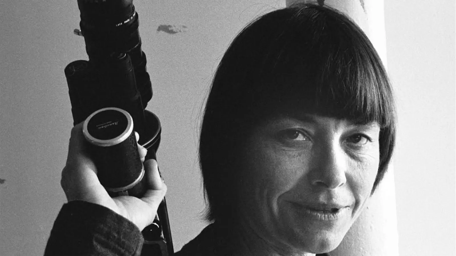 Ein schwarzweiss-Foto von der mittelalten Barbara Hammer, die selbstbewusst in den Fotoapparat blickt eine analoge Filmkamera kraftvoll in ihrer Hand und über ihrer Schulter hält.