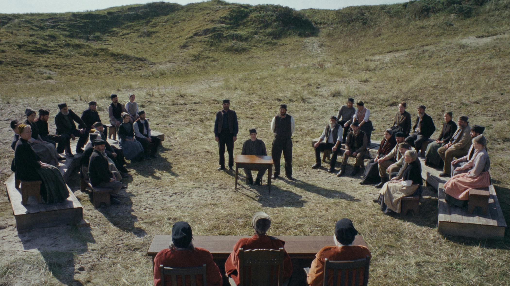 Filmstill aus DER HEIMATLOSE: Eine Dorfversammlung, genauer, das Dorfgericht, in karger Dünenlandschaft. Die Gemeinschaft sitzt auf einfachen Holzbänken in zwei Reihen einander gegenüber, im Zentrum steht ein Tisch mit einem Stuhl auf dem ein Mann (Hein) sitzt, hinter ihm zwei Wächter stehend. Vor dem Ensemble präsidiert das Gericht mit drei Richtern in roten Samtroben. Keine Gebäude, nur Sand und Gras. Foto: IFB/Florian Mag.