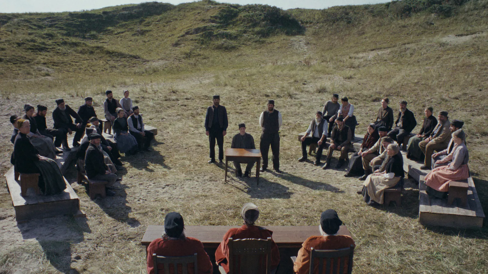 Filmstill aus DER HEIMATLOSE: Eine Dorfversammlung, genauer, das Dorfgericht, in karger Dünenlandschaft. Die Gemeinschaft sitzt auf einfachen Holzbänken in zwei Reihen einander gegenüber, im Zentrum steht ein Tisch mit einem Stuhl auf dem ein Mann (Hein) sitzt, hinter ihm zwei Wächter stehend. Vor dem Ensemble präsidiert das Gericht mit drei Richtern in roten Samtroben. Keine Gebäude, nur Sand und Gras. Foto: IFB/Florian Mag.