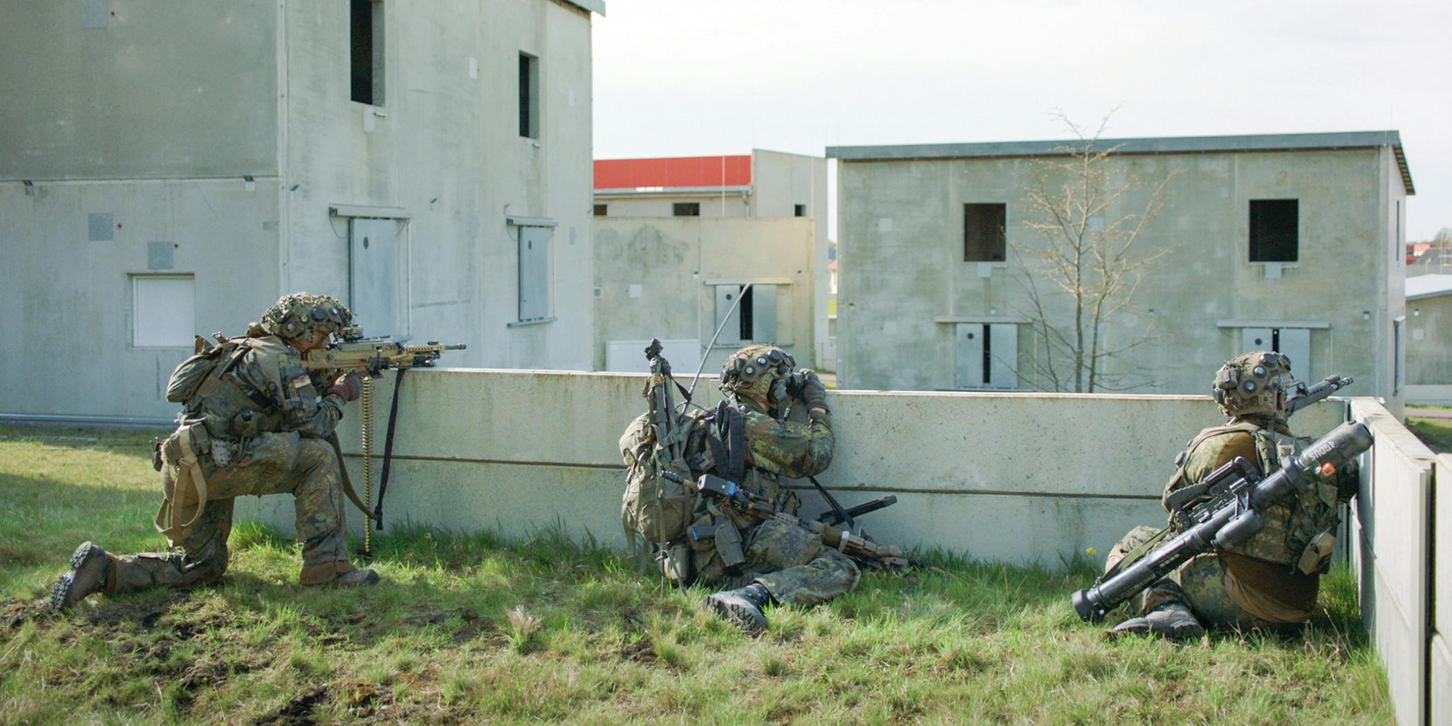 Drei Bundeswehrsoldaten in Kampfausrüstung hocken mit Gewehren in Deckung hinter einer niedrigen Betonmauer. Sie tragen Tarnuniformen und Nachtsichtgeräte. Im Hintergrund stehen mehrere kantige Betongebäude unterschiedlicher Höhe – die künstliche Übungsstadt Schnöggersburg. Filmstill aus SZENARIO von Marie Wilke.