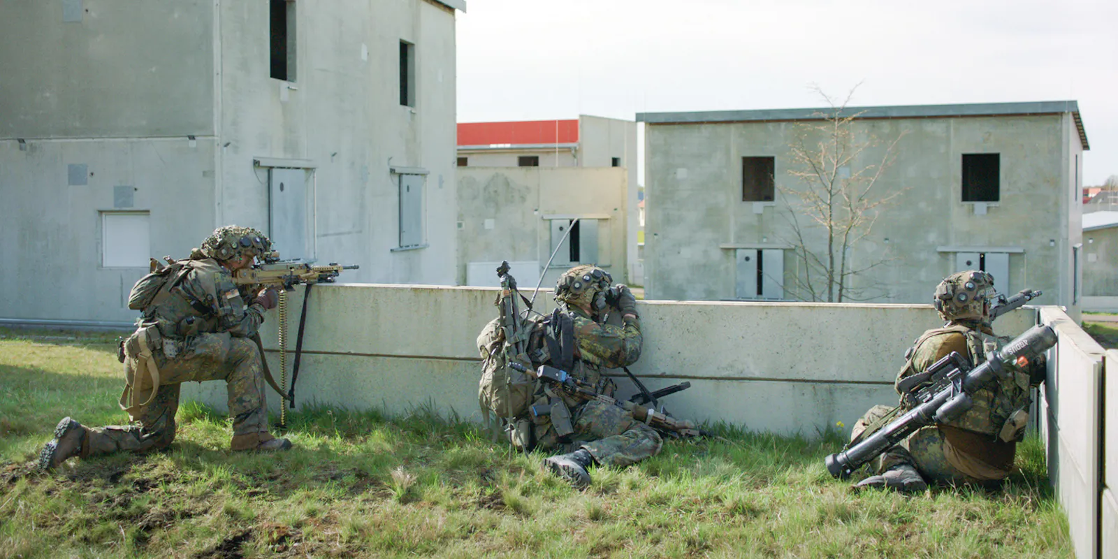 Drei Bundeswehrsoldaten in Kampfausrüstung hocken mit Gewehren in Deckung hinter einer niedrigen Betonmauer. Sie tragen Tarnuniformen und Nachtsichtgeräte. Im Hintergrund stehen mehrere kantige Betongebäude unterschiedlicher Höhe – die künstliche Übungsstadt Schnöggersburg. Filmstill aus SZENARIO von Marie Wilke.