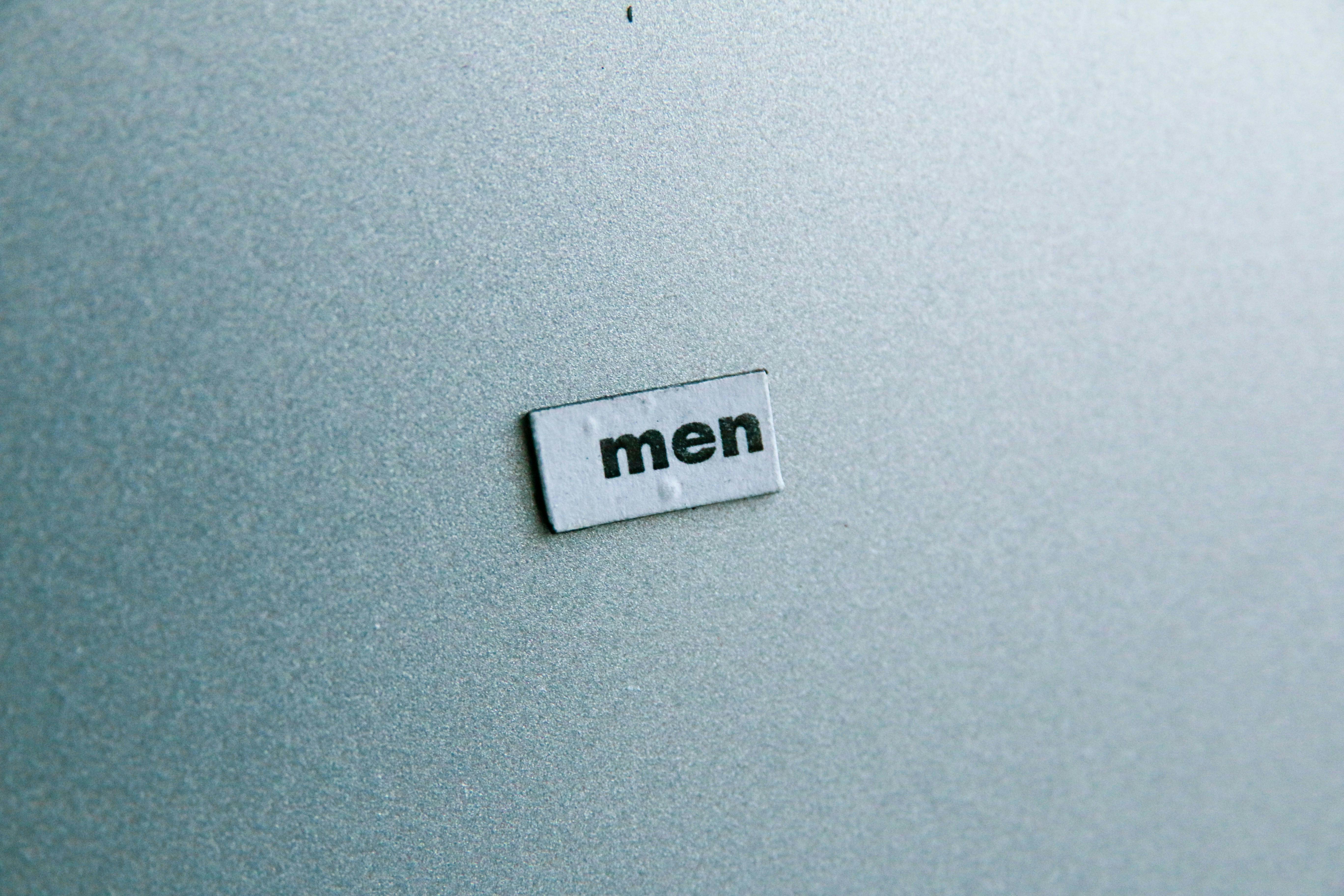 Das Wort "men" (dt.: Männer) auf einem kleinen Schild auf grauem Grund.