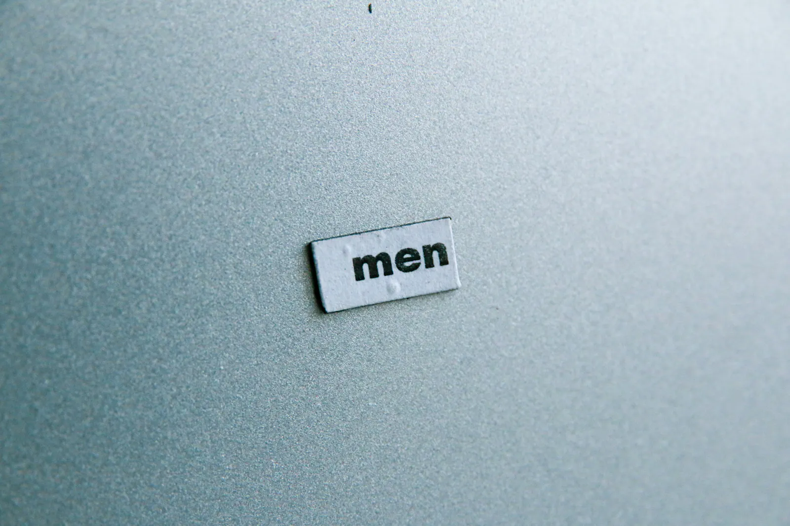 Das Wort "men" (dt.: Männer) auf einem kleinen Schild auf grauem Grund.