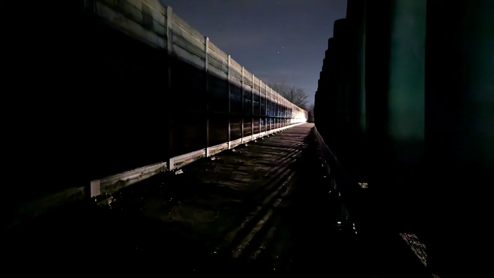 Ein Fußgängerweg im Dunkeln. Autoscheinwerfer scheinen durch Barken hindurch und werfen ein Licht auf Weg und angrenzende Lärmschutzwand, das gleichzeitig schön und gespenstisch aussieht. Am dunklen Himmel darüber leuchten vereinzelt Sterne.