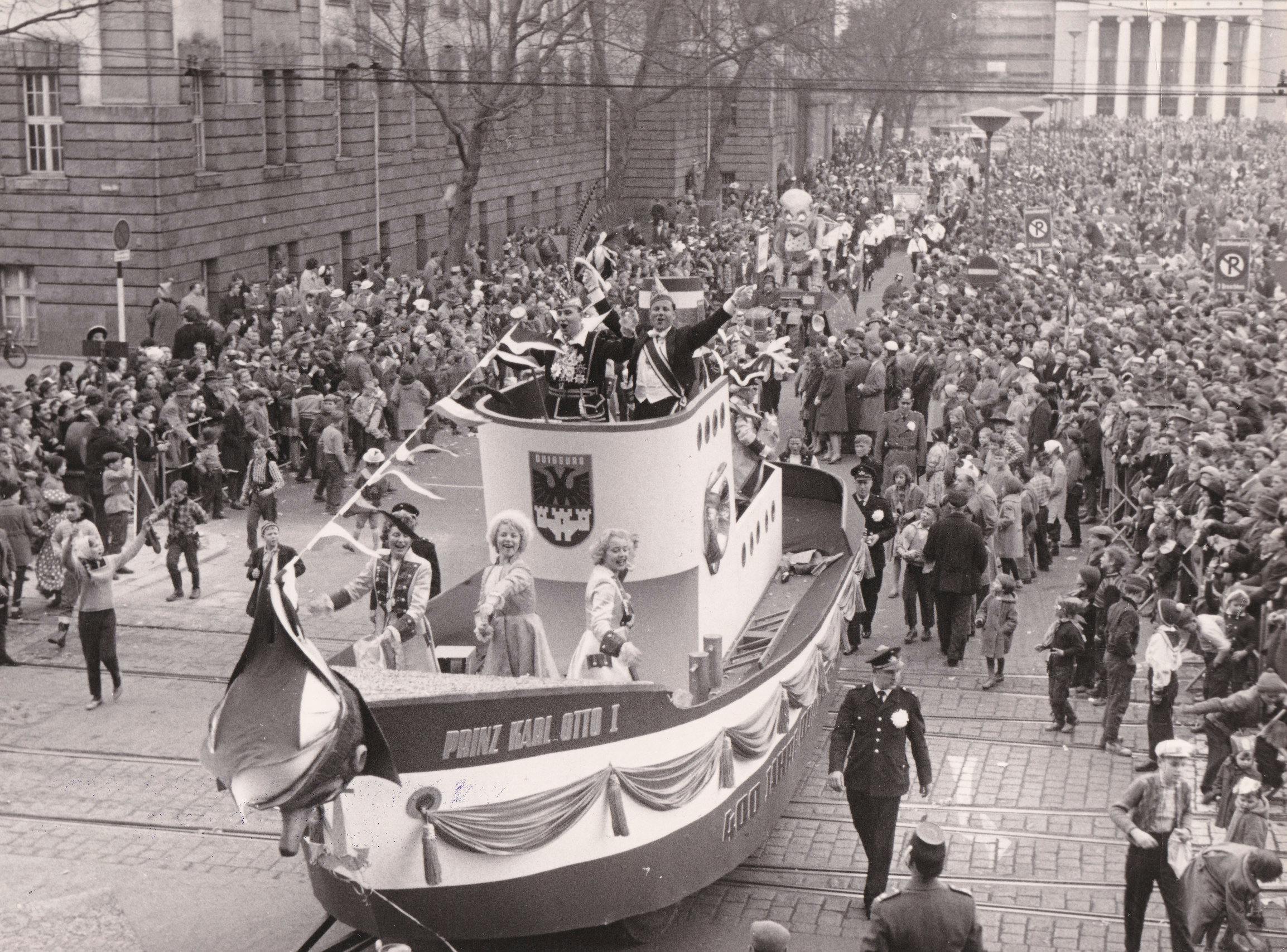 Schwarz-Weiß-Foto von einem Karnevalsumzug mit Wägen und vielen Menschen, die den Zug zuschauen. Im Vordergrund ein Wagen in Form eines Bootes mit dem Wappen der Stadt Duisburg.