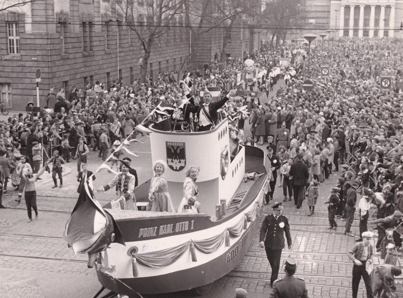Schwarz-Weiß-Foto von einem Karnevalsumzug mit Wägen und vielen Menschen, die den Zug zuschauen. Im Vordergrund ein Wagen in Form eines Bootes mit dem Wappen der Stadt Duisburg.
