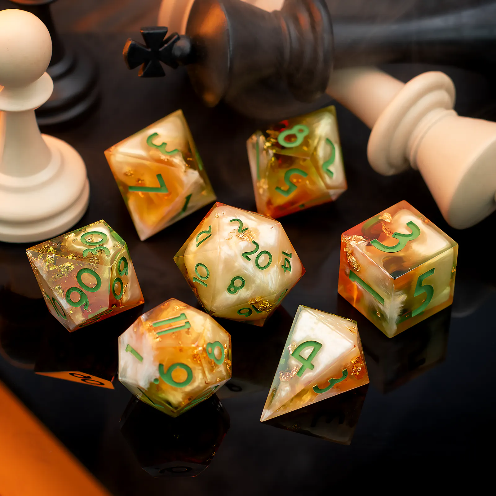 Ein siebenteiliges Würfelset aus Kunstharz in warmem Gold und Creme mit eingeschlossenen Goldfolienflocken und grüner Schrift. Die Würfel liegen auf einer spiegelnden schwarzen Oberfläche, umgeben von umgekippten Schachfiguren in Schwarz und Weiß.