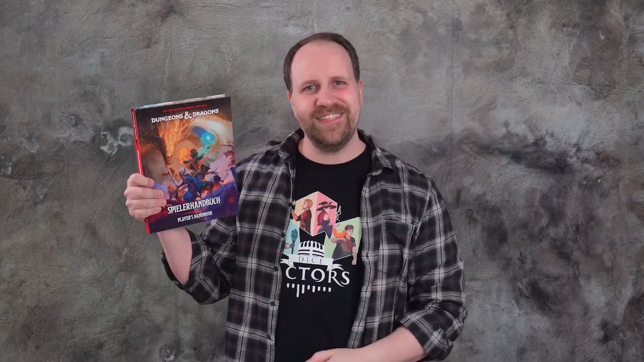 Alex steht vor einer grauen Wand, lächelt und hält das neue Spielerhandbuch von Dungeons & Dragons in die Kamera.