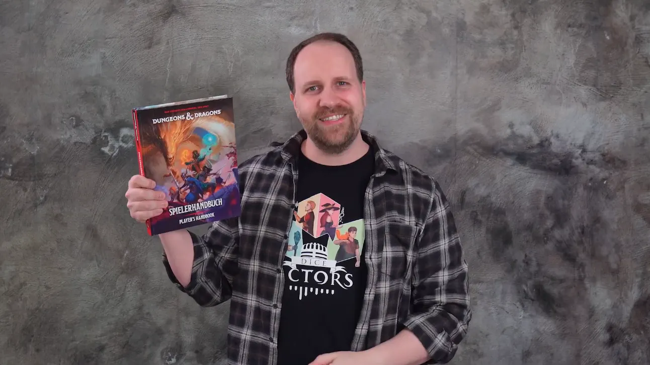 Alex steht vor einer grauen Wand, lächelt und hält das neue Spielerhandbuch von Dungeons & Dragons in die Kamera.