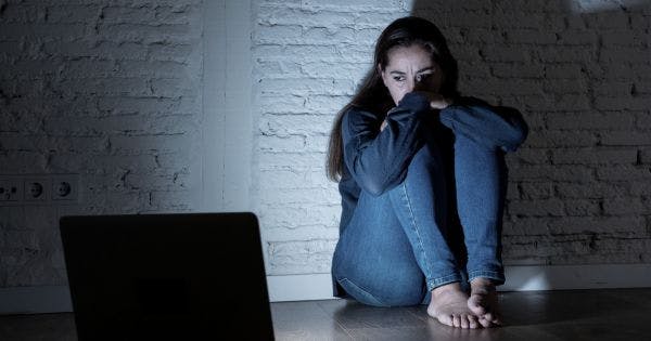 Eine junge Frau sitzt im Dunkeln an der Wand mit verzweifeltem Gesichtsausdruck. Sie starrt auf ihren Laptop. / Bild: Canva