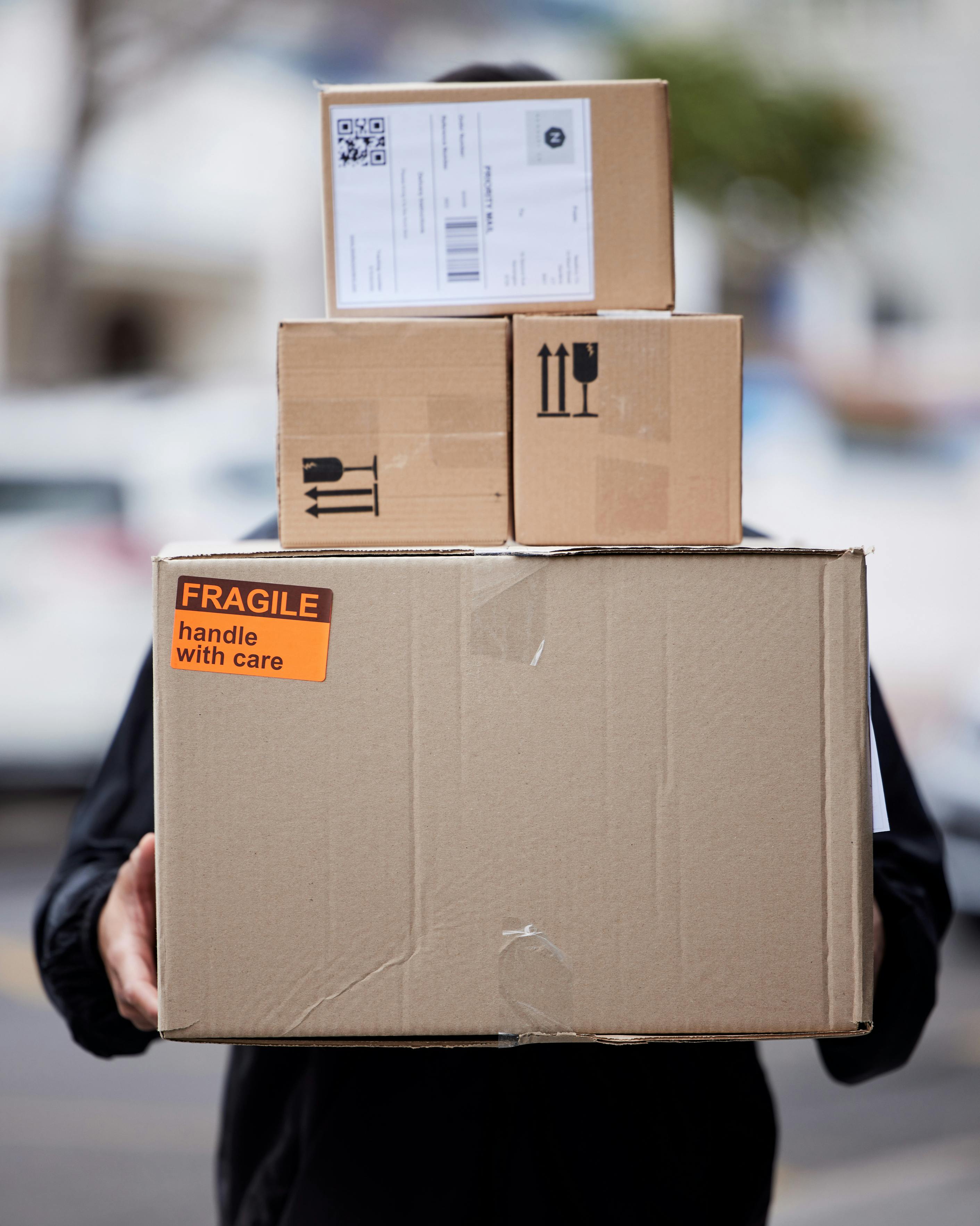 Das Bild zeigt eine Person in dunkler Kleidung, die drei übereinandergestapelte Pakete trägt. Auf dem untersten Paket klebt ein orangefarbener Aufkleber mit der Aufschrift „Fragile – handle with care".