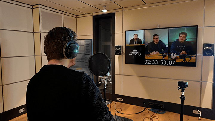 Tim steht im Tonstudio der Arena Synchron GmbH. Er wurde von hinten fotografiert. Er trägt Kopfhörer und steht vor einem Mikrofon mit Popschutz. Auf einem Fernseher an der Wand ist eine Episode der Dice Actors mit einem eingeblendeten Timecode (02:33:53:07) zu sehen. Links von Tim ist ein weiterer Bildschirm mit einem offenen englischen Dialogbuch.