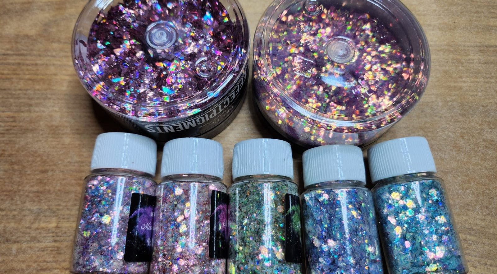 Mehrere transparente Behälter und kleine Fläschchen mit schillerndem Glitzer in verschiedenen Farben liegen auf einem Holztisch, der Glitzer reflektiert buntes Licht.