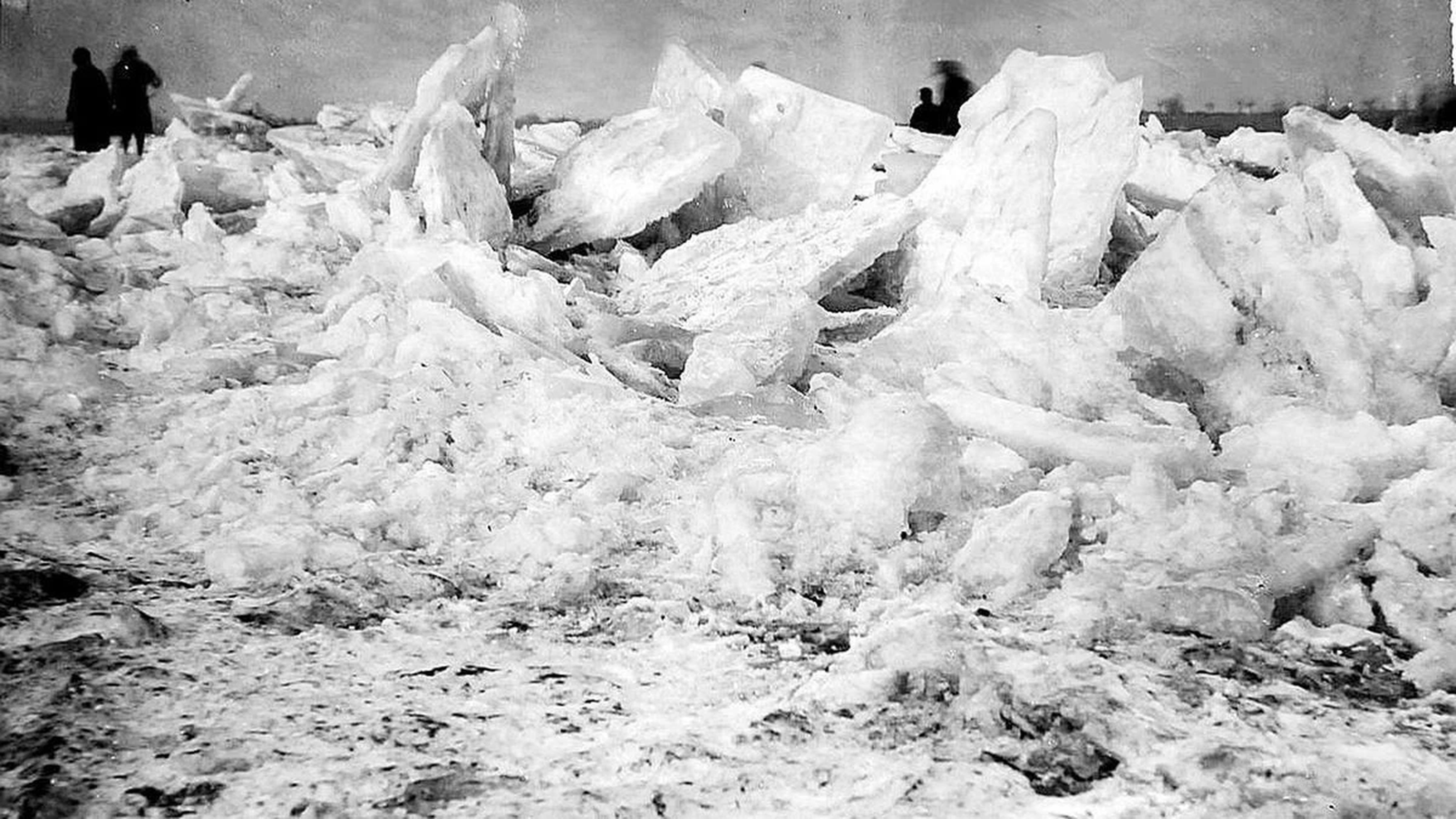 Auftürmende Eisschollen des zugefrorenen Rheins. Schwarz-Weiß. Im Hintergrund Menschen, die als schwarze kleine Umrandungen am oberen Bildrand zu sehen sind. Mutmaßlich Rhein bei Duisburg 1920er Jahre (Foto: Stadtarchiv Duisburg).