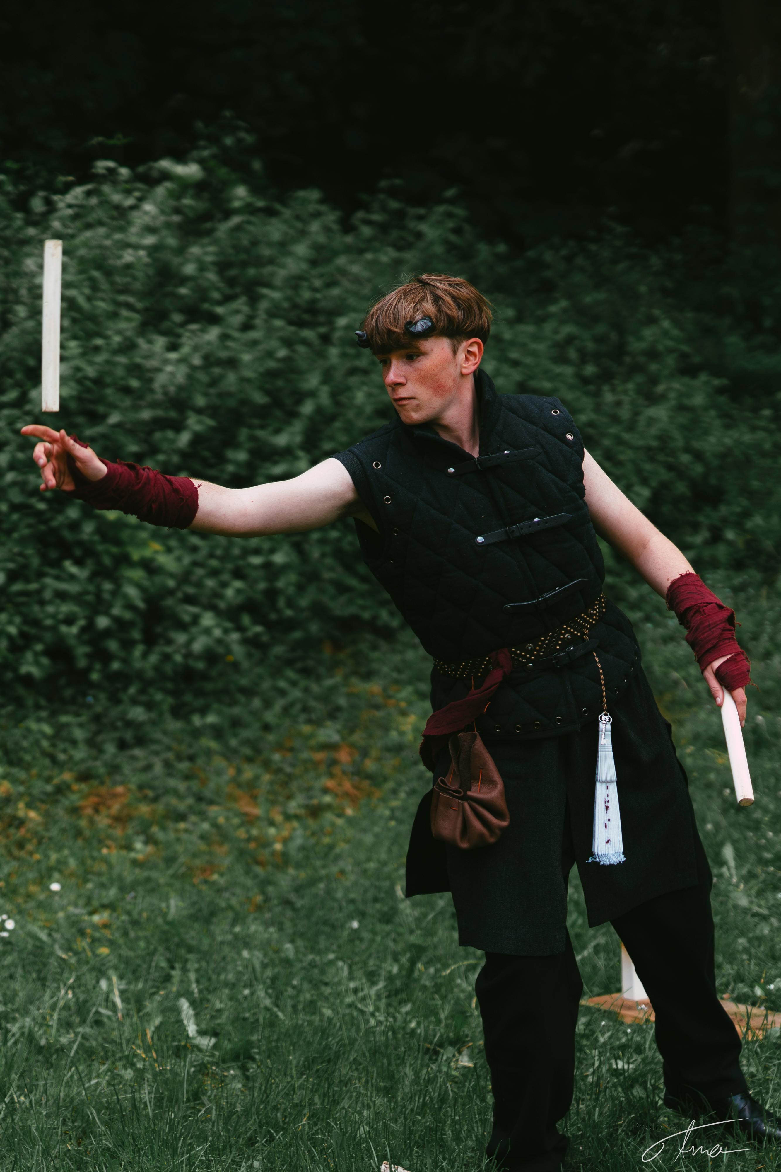Ein LARPer trägt eine dunkle Weste und eine dunkle Hose. Seine Hände sind, einem Mönch ähnlich, praktisch mit rotem Stoff umwickelt. Zwei kurze schwarze Hörner kommen aus seinen Schläfen. Er wirft einen kleinen Stock, steht im grünen und scheint ein Wurfspiel zu spielen.