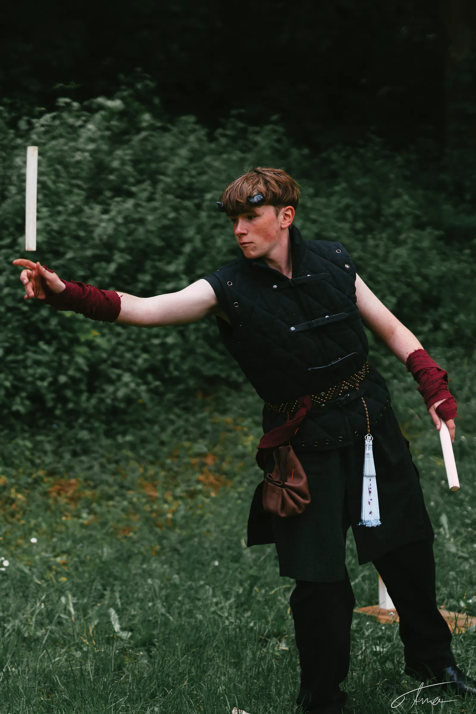 Ein LARPer trägt eine dunkle Weste und eine dunkle Hose. Seine Hände sind, einem Mönch ähnlich, praktisch mit rotem Stoff umwickelt. Zwei kurze schwarze Hörner kommen aus seinen Schläfen. Er wirft einen kleinen Stock, steht im grünen und scheint ein Wurfspiel zu spielen.