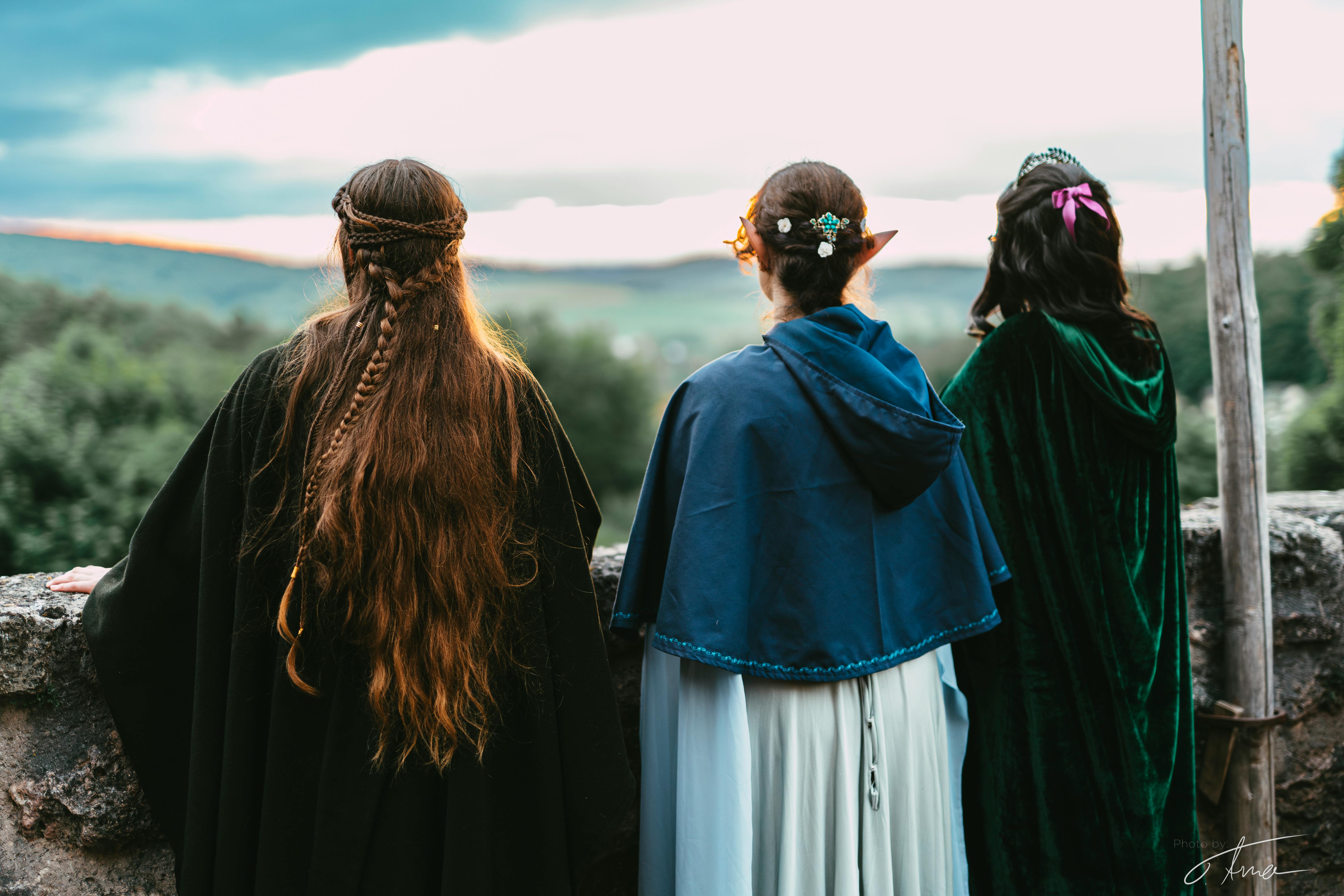 Drei verkleidete LARP-Spielende sind von hinten zu sehen. Sie schauen an einer Burgmauer hinaus in den Sonnenuntergang. Links ist eine Frau mit dunklem Umhang und langen, rotbraunen Haaren. In der Mitte eine Elfe mit bläulichem Umhang, Dutt und Schmuck in den Haaren und rechts eine Frau mit dunklen Haaren, einer pinken Schleife und einem samtenen grünen Umhang.