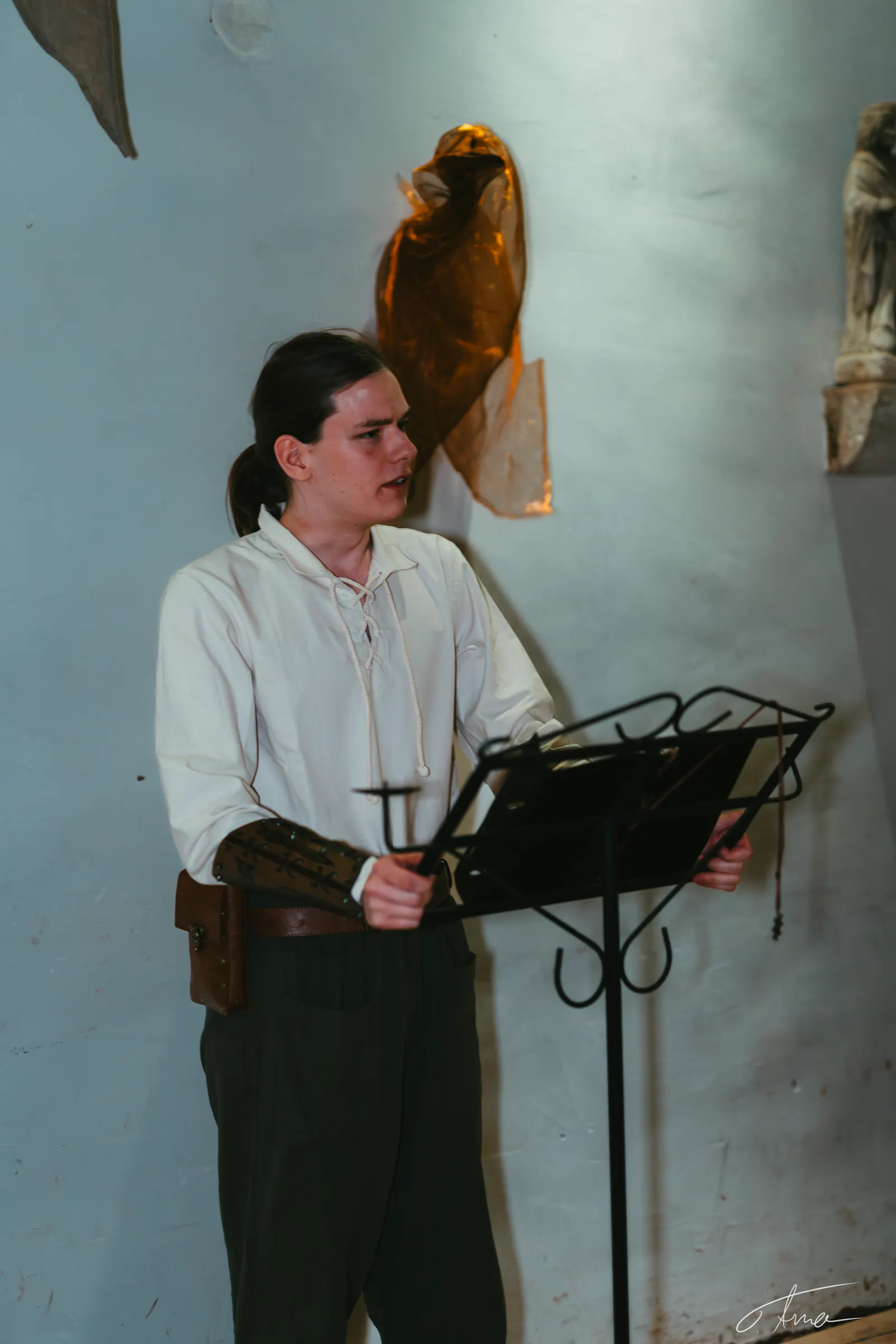 Ein LARPer steht an einem Pult und hält eine Rede vor einer weißen Wand. Er trägt ein einfaches weißes Leinenhemd, lederne Armschiene und eine schwarze Hose.