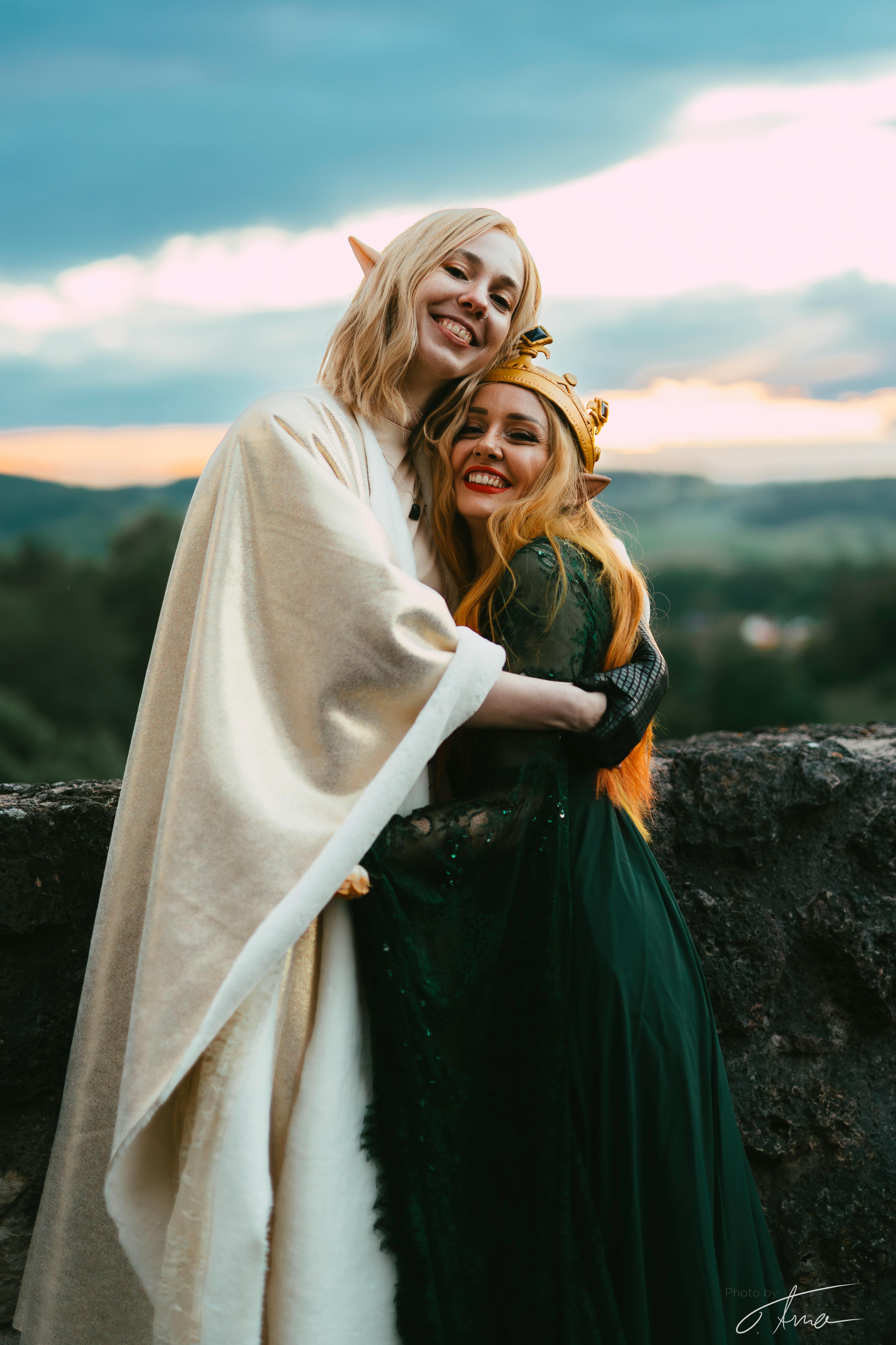Im Sonnenuntergang lächeln und umarmen sich eine Elfe mit hellblonden Haaren und weiß-goldenem Umhang sowie eine Elfe mit blonden Haaren, einer Krone und grünlichem Kleid.