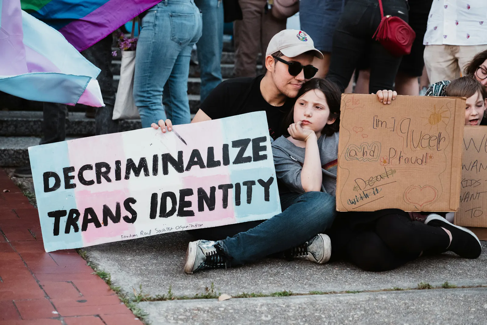 Eine Versammlung: Eine Person mit Sonnenbrille und Basecap sitzt mit einem älteren Kind auf dem Boden. Beide halten ein Schild. Auf dem blau-rosa-weiß gestreiften Schild der erwachsenen Person steht: Decriminalize Trans Identity.