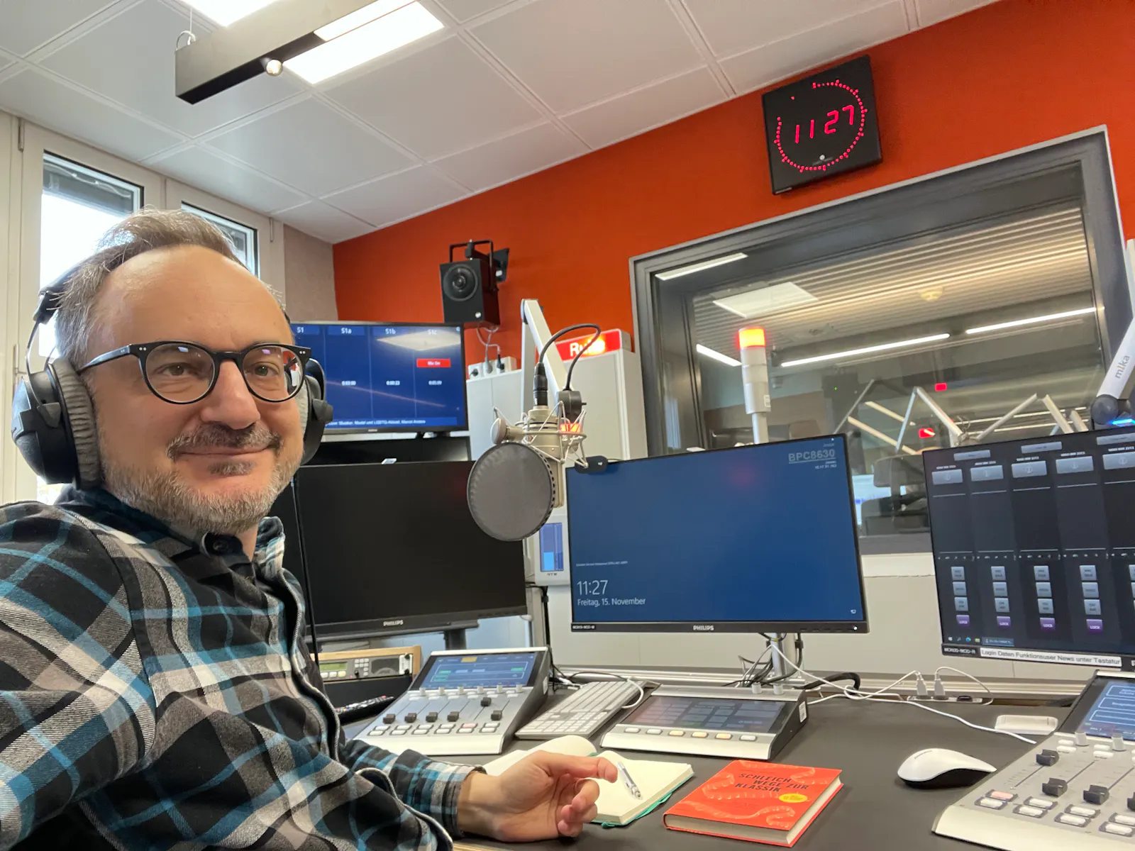 Ich sitze in einem blau-schwarz kariertem Holzfällerhemd in einem Aufnahmestudio voller Bildschirme