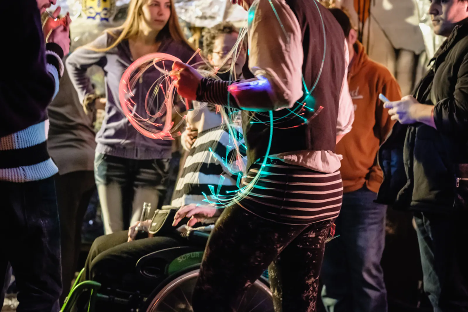 Mehrere Menschen stehen dicht beieinander, eine davon nutzt einen Rollstuhl, mehrere haben fluoreszierende Bänder um und tanzen.
