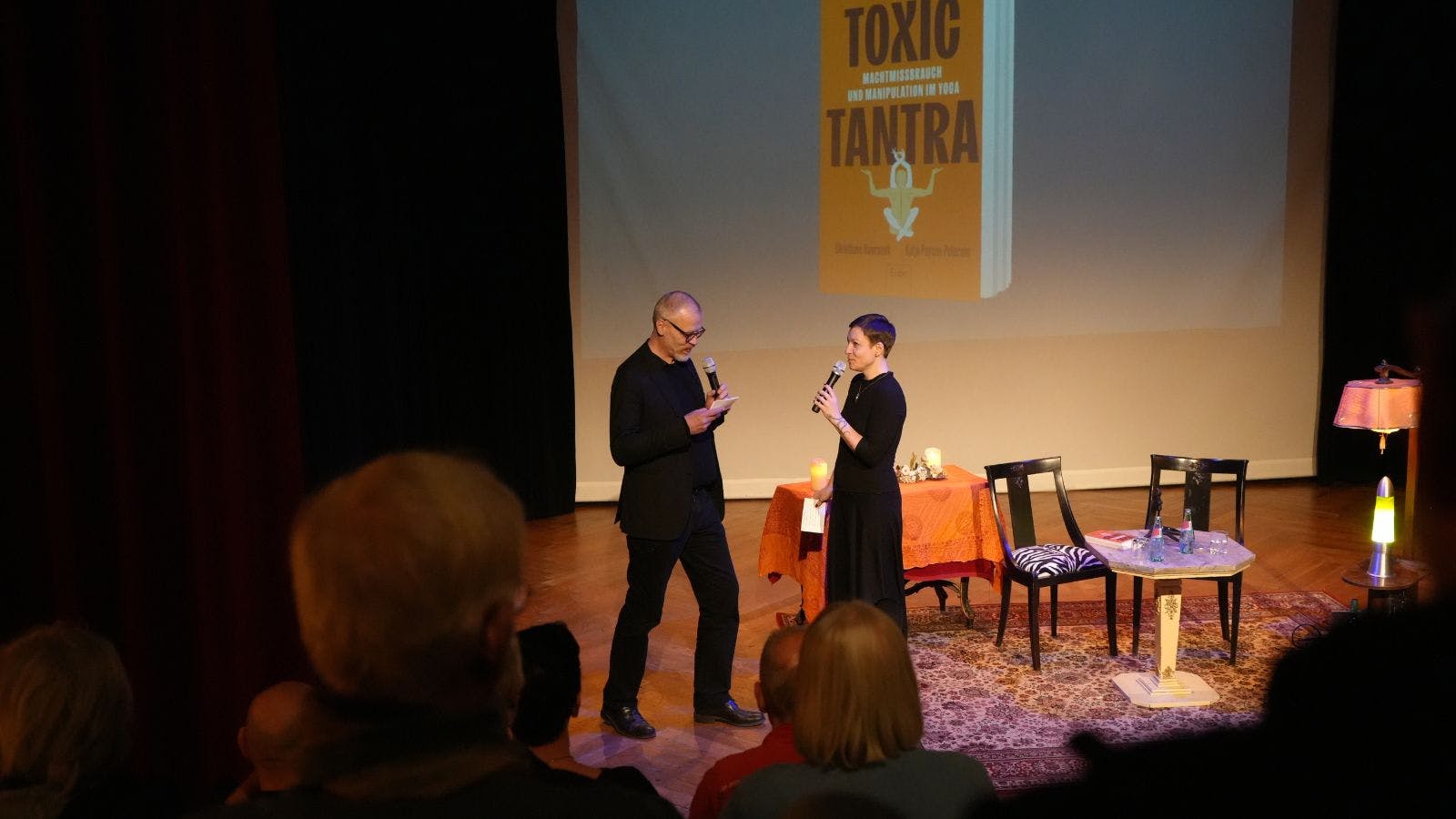 Eva Hanson und Ulf Torreck moderieren Sex, Death and Magic. Sie stehen auf einer Theaterbühne, im Hintergrund ist das Cover von "Toxic Tantra" zu sehen