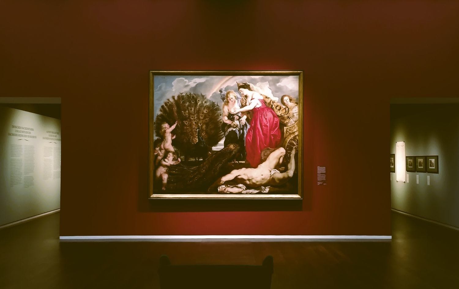 Ein großes Bild hängt an einer roten Museumswand. Auf dem Bild sieht man drei Frauen, einen Mann, der auf dem Boden liegt, links zwei Putten.