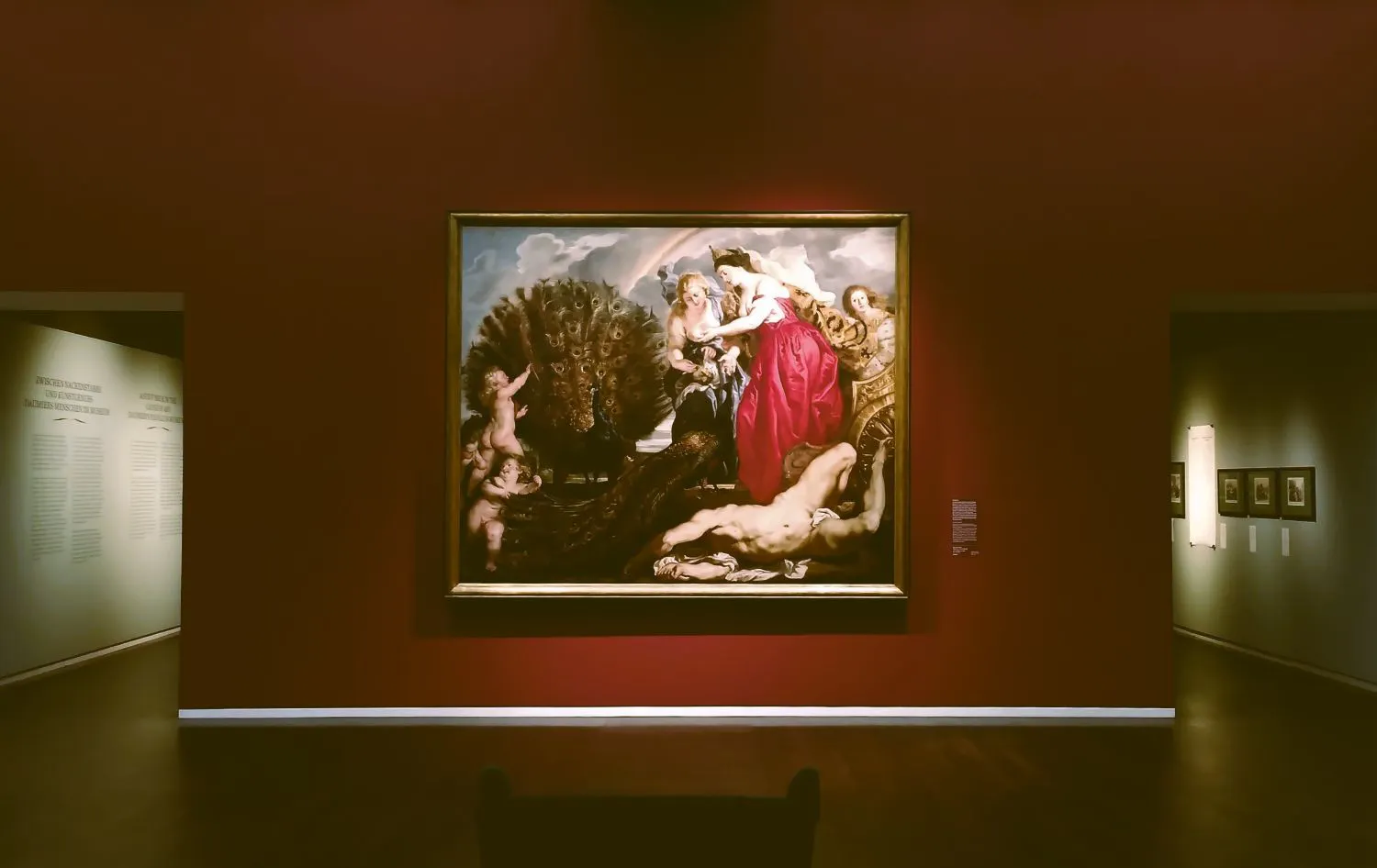 Ein großes Bild hängt an einer roten Museumswand. Auf dem Bild sieht man drei Frauen, einen Mann, der auf dem Boden liegt, links zwei Putten.