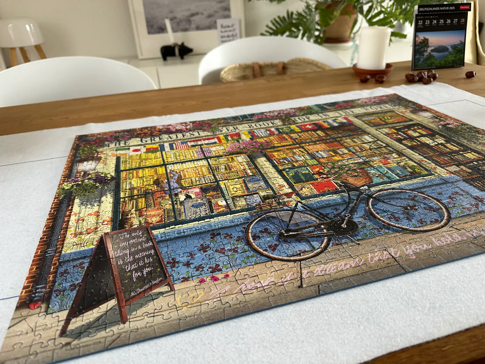Ein fertiges Puzzle auf einer weißen Puzzle-Matte auf einem Holztisch. Das Motiv: Eine Buchhandlung von draussen mit einem Fahrrad davor und einer handbeschriebenen Werbetafel.