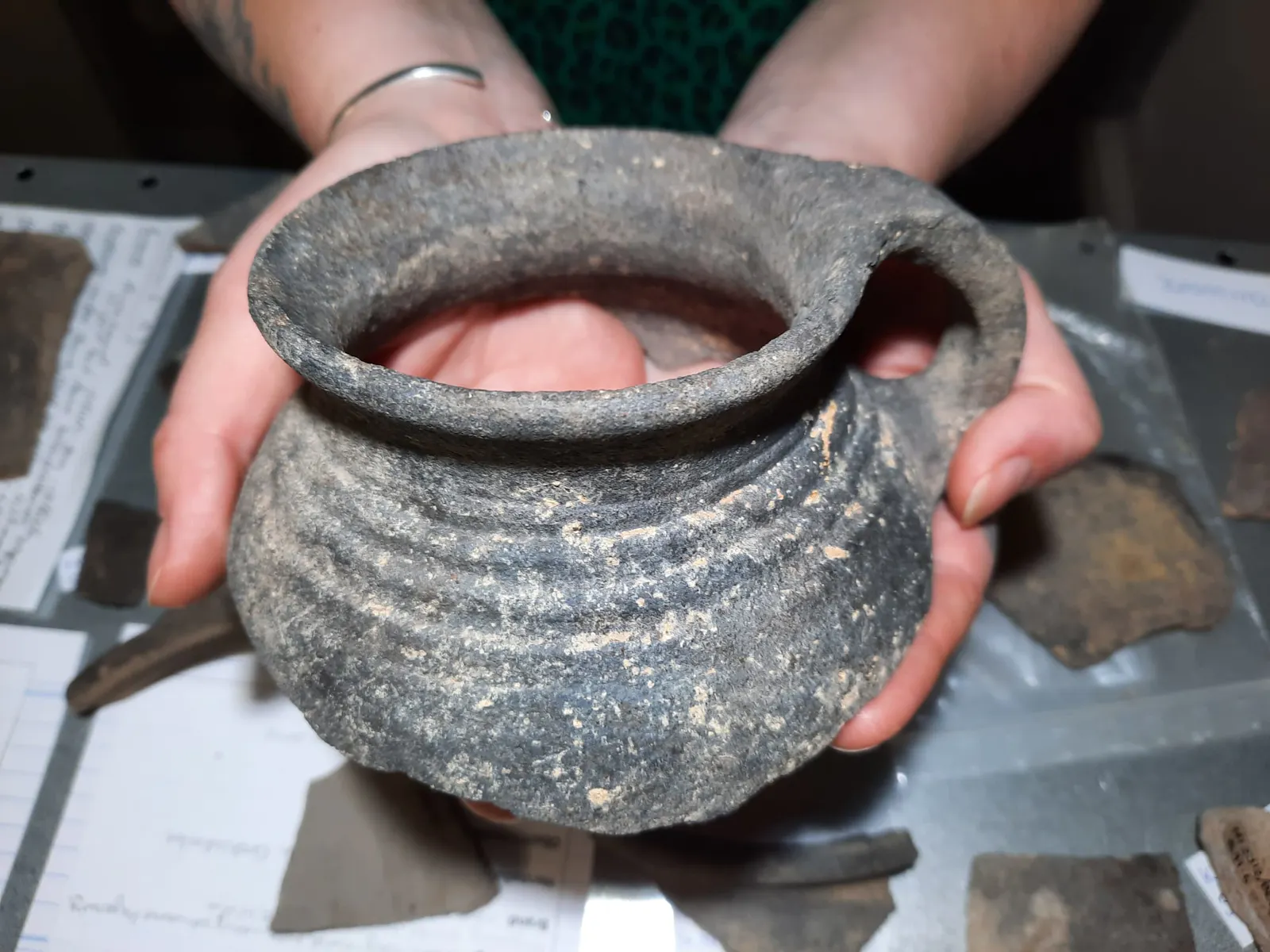 Eine Frau hält den oberen Teil eines Keramiktopfes. Die Mündung der grauen Ware ist vollständig erhalten und der Topf hat einen Henkel. (Foto: Karina Schnakenberg)