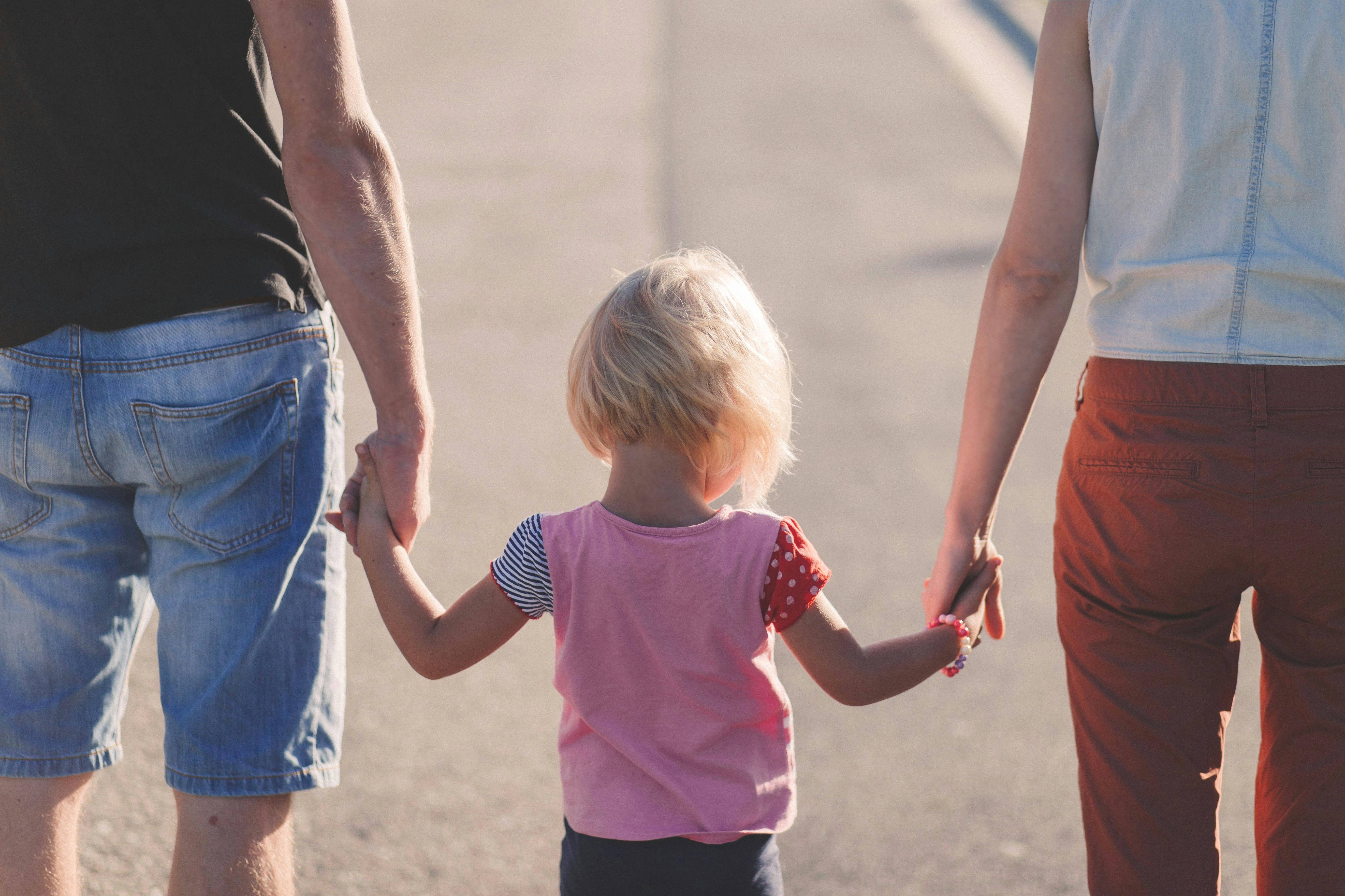 Ein junges Mädchen mit kurzen blonden Haaren und einem rosa T-Shirt hält die Hände seiner Eltern. Wir blicken von hinten auf die Szene und sehen ihre Rücken. Der Vater trägt eine kurze Jeans, die Mutter eine rote Hose. Sie laufen auf einer Straße.