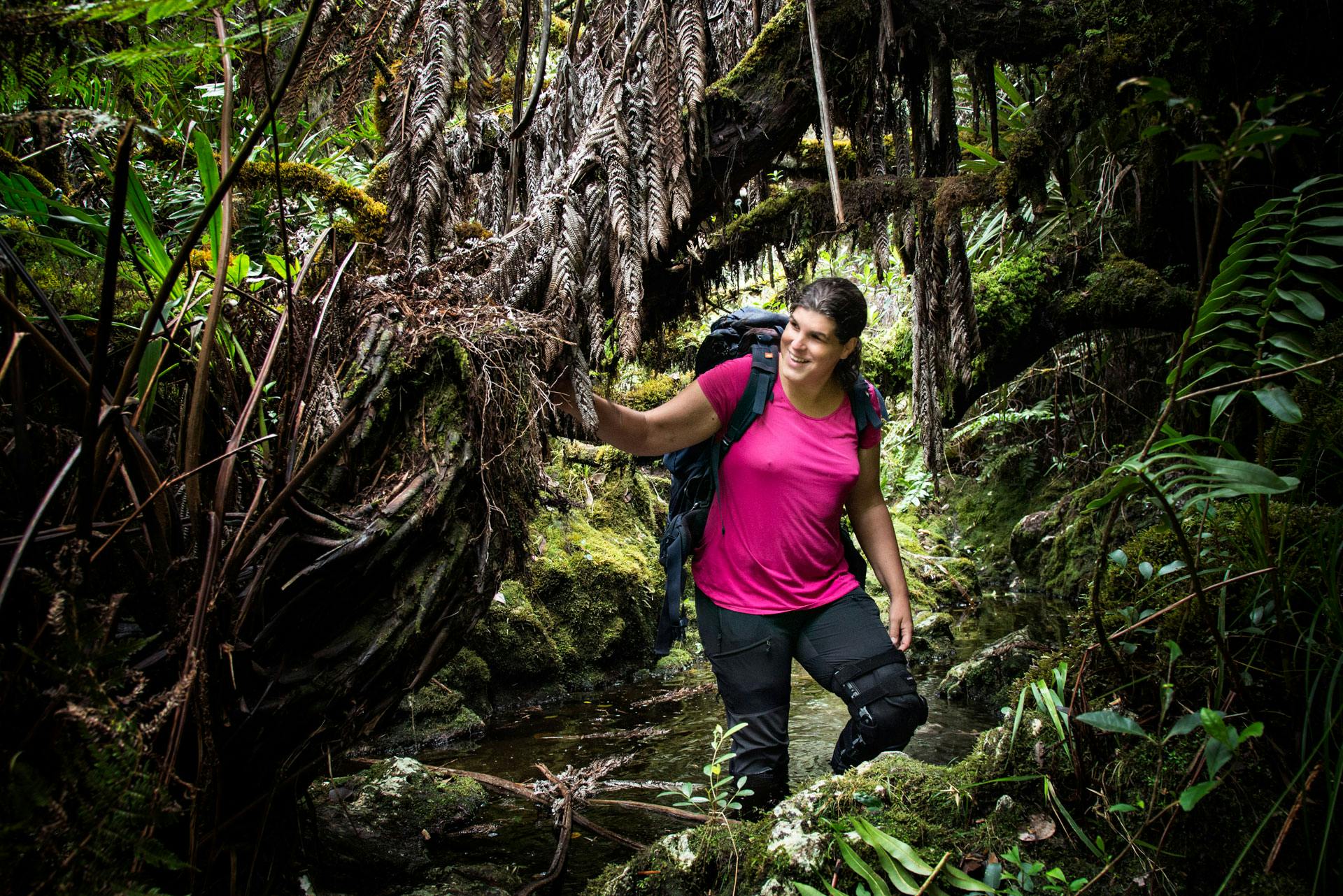 Diane en vêtements techniques et avec un gros sac à dos, souriante, marchant dans un ruisseau au cœur d'une végétation tropicale luxuriante.