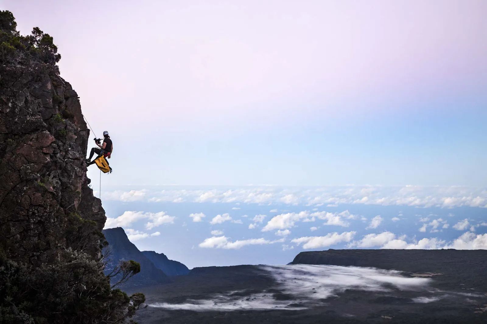 Diane suspendue en rappel contre une falaise, très haut au-dessus d'une plaine volcanique couverte de brume. À l'horizon en arrière-plan, des nuages et l'Océan Indien à perte de vue.