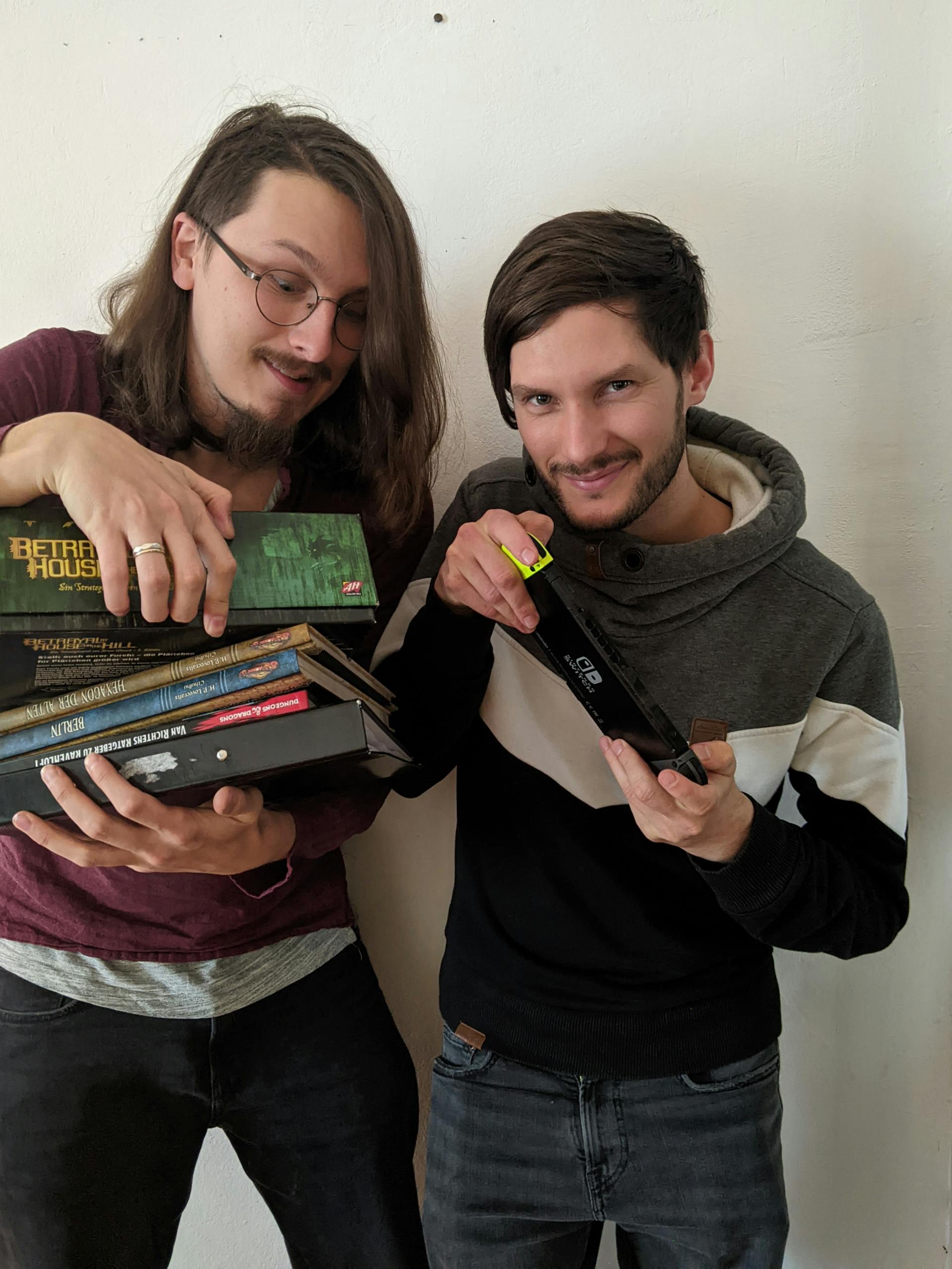 Paul und Jakob stehen vor einer weißen Wand und halten gemeinsam einen Stapel Bücher und eine Switch in den Händen. Paul steht links. Er trägt eine Brille und hat lange Haare. Jakob steht rechts neben ihm und lächelt in die Kamera.