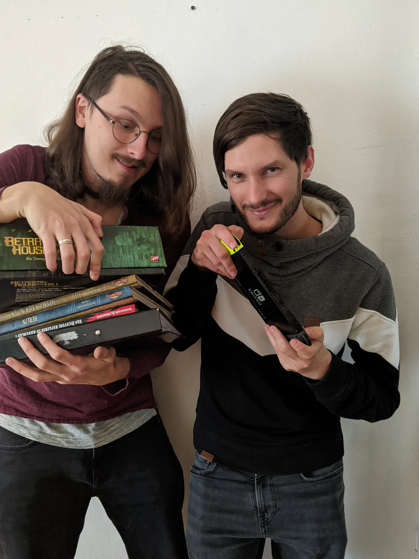 Paul und Jakob stehen vor einer weißen Wand und halten gemeinsam einen Stapel Bücher und eine Switch in den Händen. Paul steht links. Er trägt eine Brille und hat lange Haare. Jakob steht rechts neben ihm und lächelt in die Kamera.