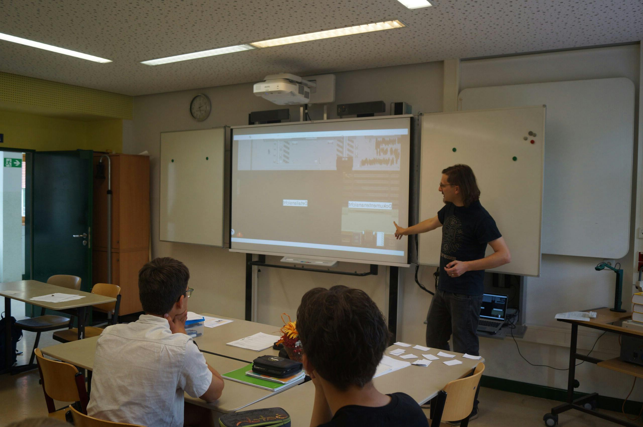 Paul steht vorne in einem Klassenraum und zeigt auf eine projizierte Präsentation an einem großen Bildschirm. Mehrere sitzende Zuhörende verfolgen den Vortrag aufmerksam an Tischen mit Unterlagen und Notizen. Der Raum ist hell beleuchtet und mit Whiteboards sowie technischer Ausstattung ausgestattet.