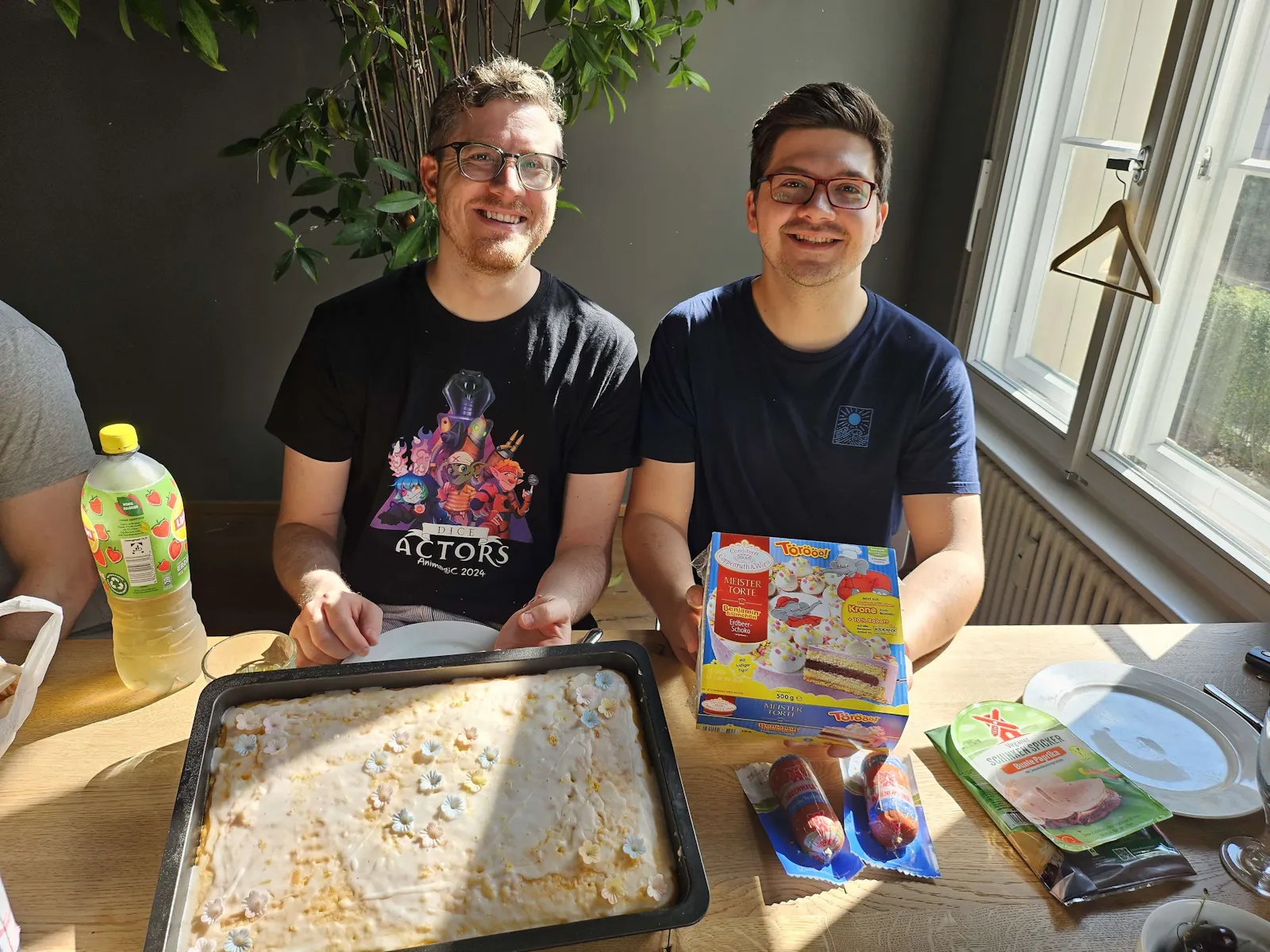 Tim und Patze sitzen mit Zitronenblechkuchen und Benjamin Blümchen-Torte am Frühstückstisch und grinsen.
